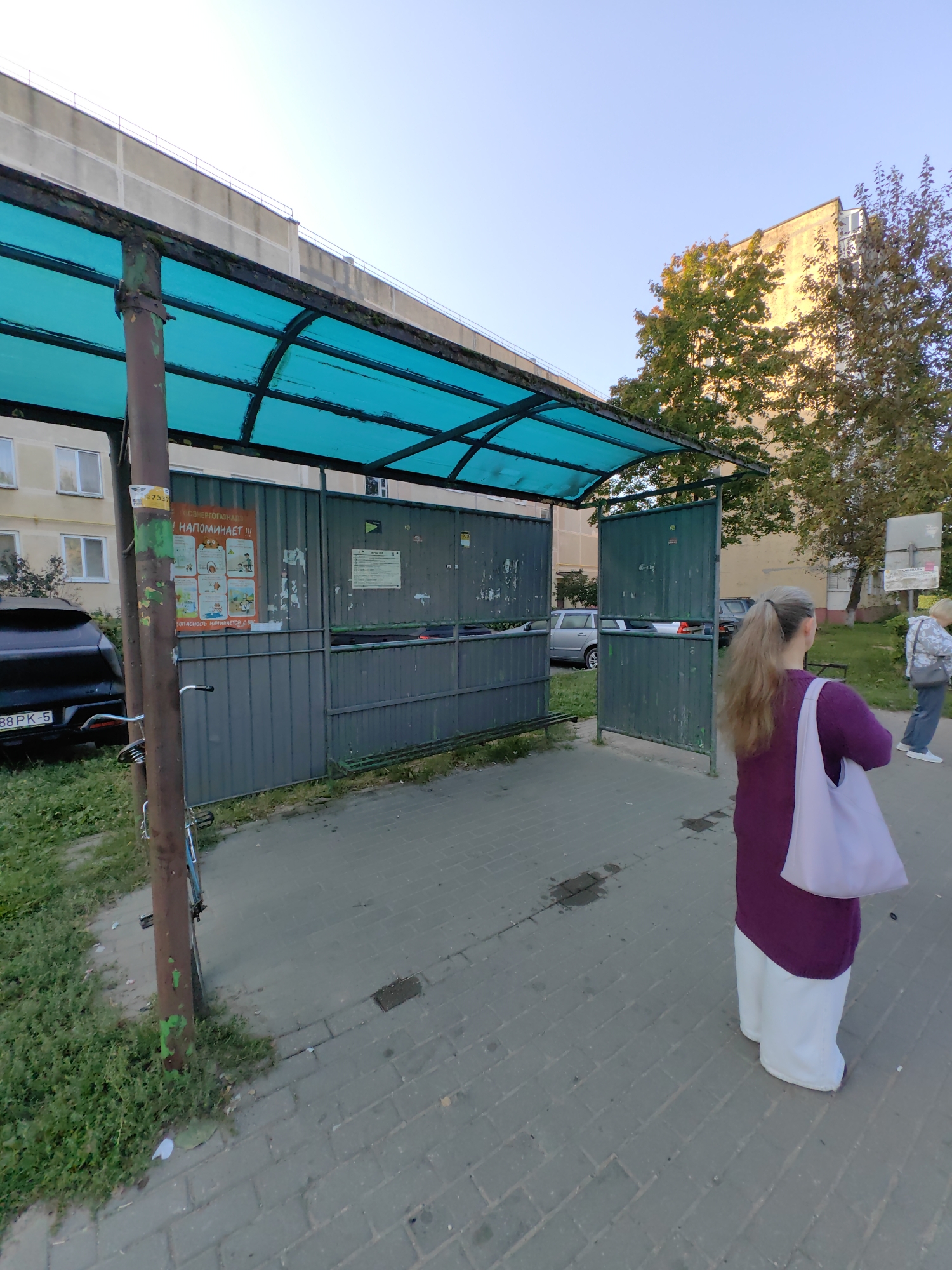 The image depicts a street scene with a bus stop shelter, a building, and several people. The bus stop shelter has a turquoise canopy and gray metal walls. A bicycle is leaning against a pole in front of the shelter. A black car with a license plate that reads "88 PK-5" is parked to the left of the shelter. In the background, a multi-story building can be seen.
A woman with blonde hair in a ponytail is standing in the foreground, facing away from the camera, wearing a purple top and white pants, carrying a white tote bag. To the right, another woman in a light-colored jacket and dark pants is walking away from the camera.
The scene appears to be in Fanipal, Belarus, as indicated by a sign on the bus stop shelter. The environment is urban, with paved walkways and grassy areas. The sky is clear and blue. The time of day appears to be late afternoon or early evening, given the lighting and the long shadows.