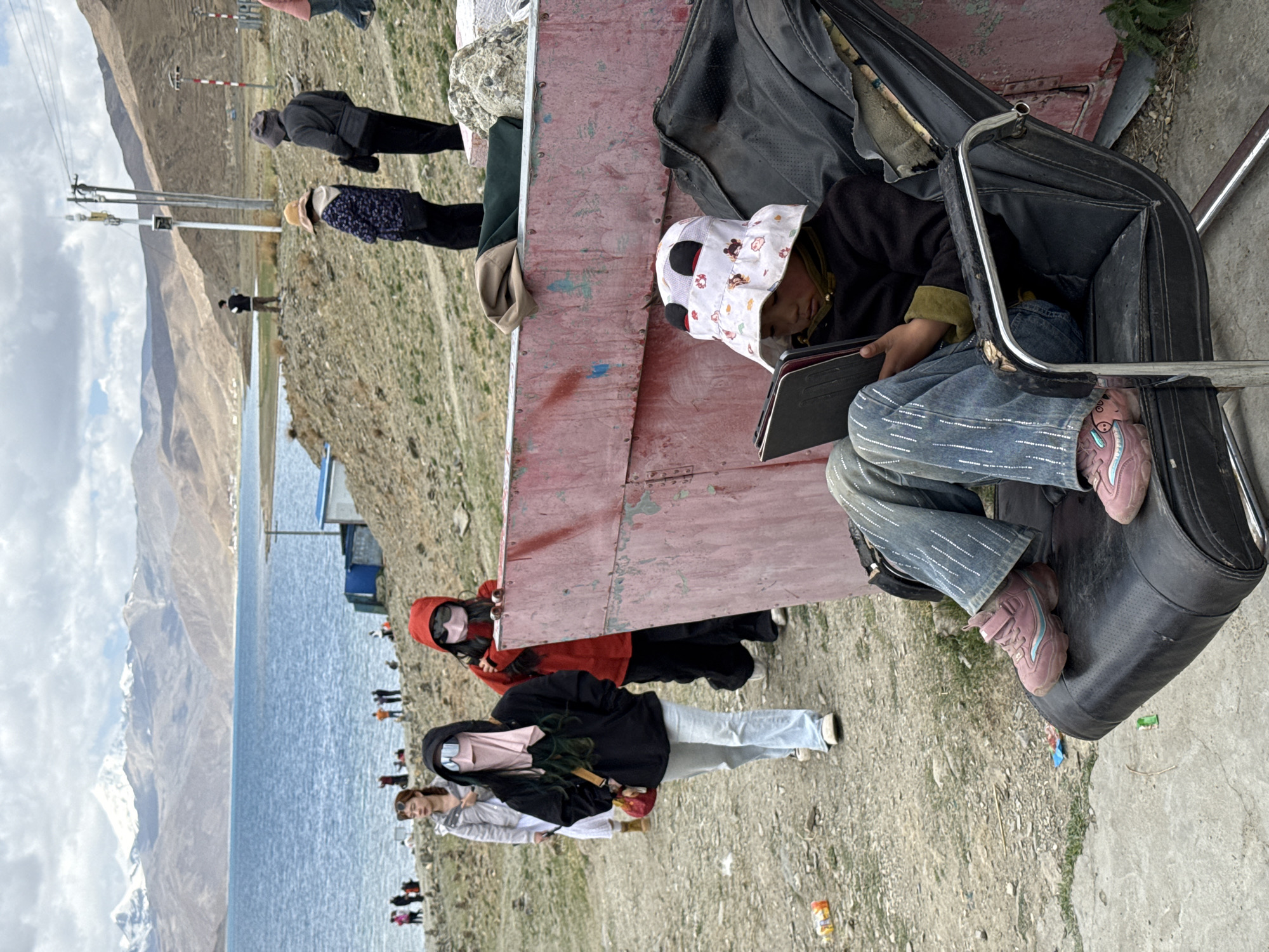 A lakeside landscape at Baidê, China, features a girl in a panda-themed sunhat and denim, absorbed in a tablet while seated on a black vehicle accessory near a weathered red structure. Three women with concealed faces stand nearby, overlooking the lake, where other visitors are visible. Mountains are in the background under a cloudy sky.