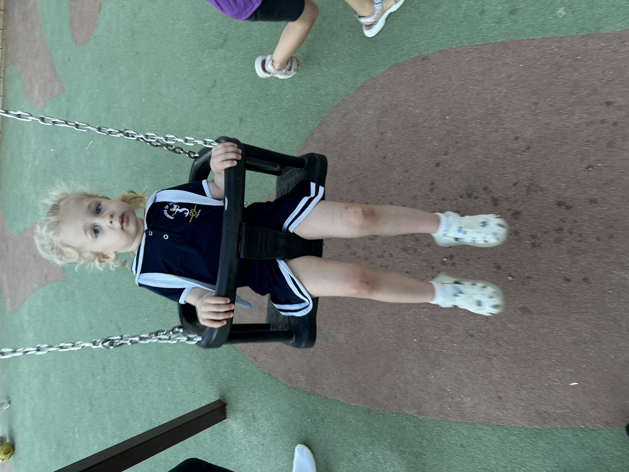 A young blonde child with blue eyes is seated on a swing in a playground. The child is wearing a navy blue shirt with white trim and a white collar, and navy blue shorts with white stripes. They have white socks and white patterned Croc-style shoes. The swing set has black plastic seats and metal chains. The playground surface is a mix of green and brown material, possibly rubberized safety surfacing. Another child, wearing a purple shirt and black shorts, is visible in the background on the right, their legs and feet in motion. A yellow ball is partially visible in the upper left corner. The image appears to be taken during daytime. The location is the playground of the Parc del Llastres, situated in the city of Sant Feliu de Guíxols, Spain, which is a coastal town in Catalonia, Spain.