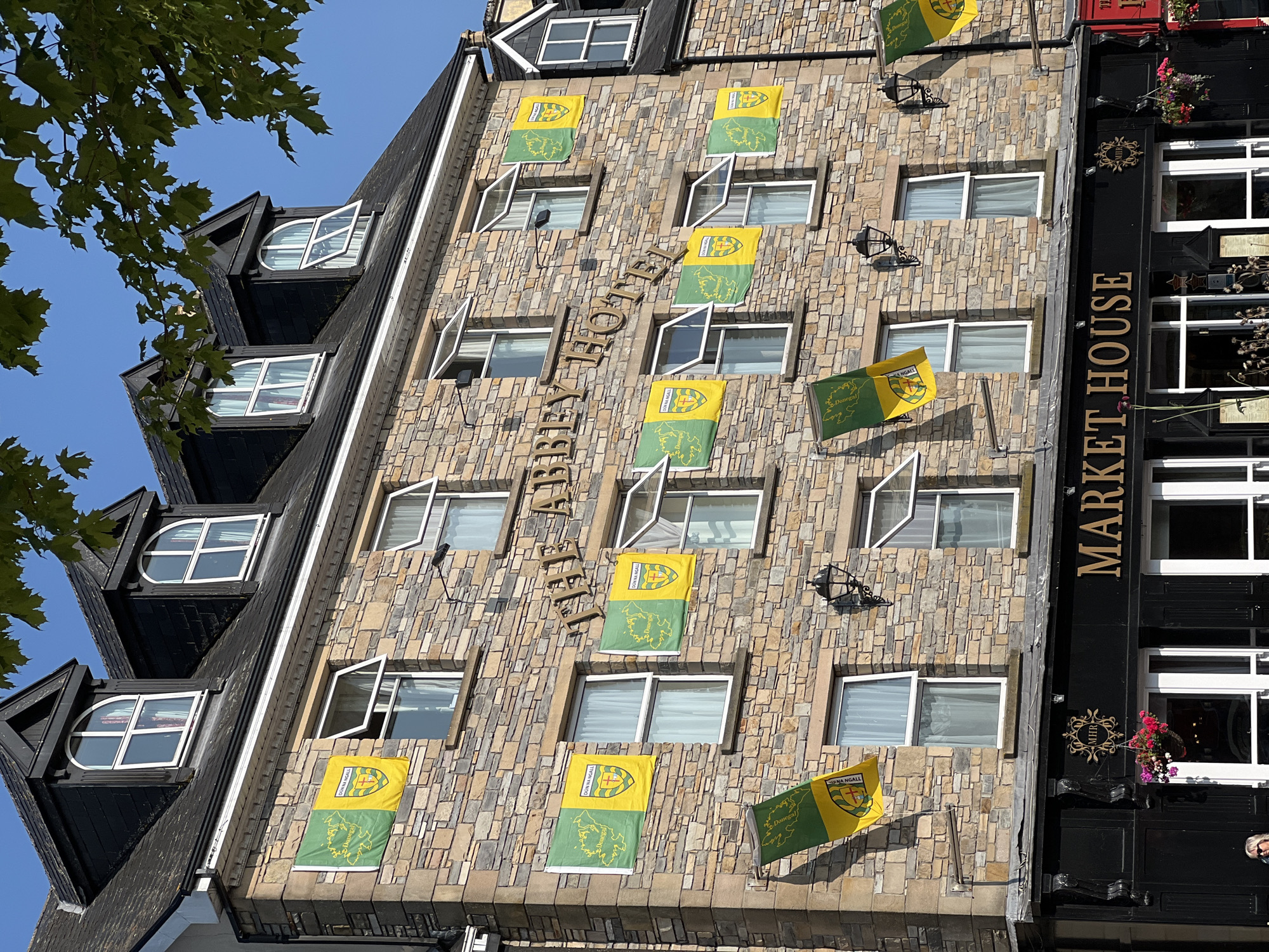 The image shows the facade of "The Abbey Hotel" located in the city of Donegal, Ireland. The building is constructed of stone with multiple stories and dormer windows. Numerous flags, predominantly green and yellow with a crest, are displayed on the facade, suggesting a local or sporting affiliation. The name "The Abbey Hotel" is visible in large, gold lettering above the flags. Below the hotel is a sign that reads "Market House". The scene is captured during daylight with a clear blue sky. Foliage from a tree hangs into the upper portion of the frame.