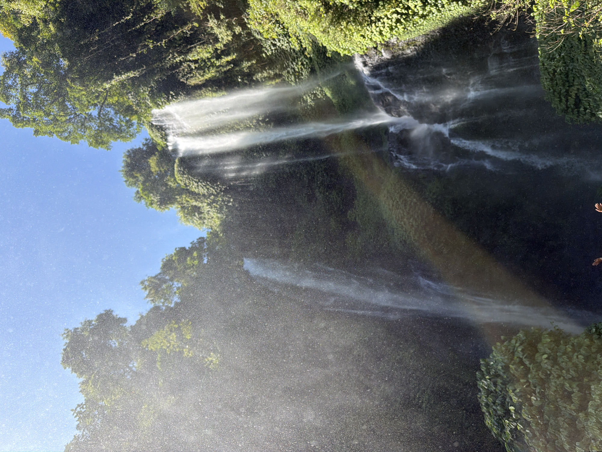 In the city of Sawan, Indonesia, a tall waterfall cascades down a lush, green cliff face. Sunlight streams through the canopy of trees, illuminating the falling water and creating a misty spray. The water plunges into a pool below, surrounded by dense vegetation and moss-covered rocks. The scene is vibrant and serene, capturing the natural beauty of the tropical landscape.