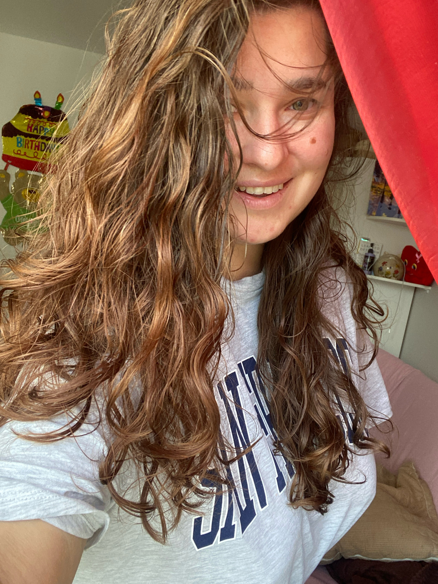 The image shows a young woman indoors, smiling at the camera. She has long, wavy brown hair that partially obscures her face. She is wearing a light-colored t-shirt with dark blue lettering. Behind her, a red curtain is visible, as is a portion of a wall with a birthday balloon shaped like a cake and some shelves with decorative objects. The background suggests a casual, domestic setting, possibly a bedroom or living room. There is no discernible information indicating the location in the city of Perm, Russia.