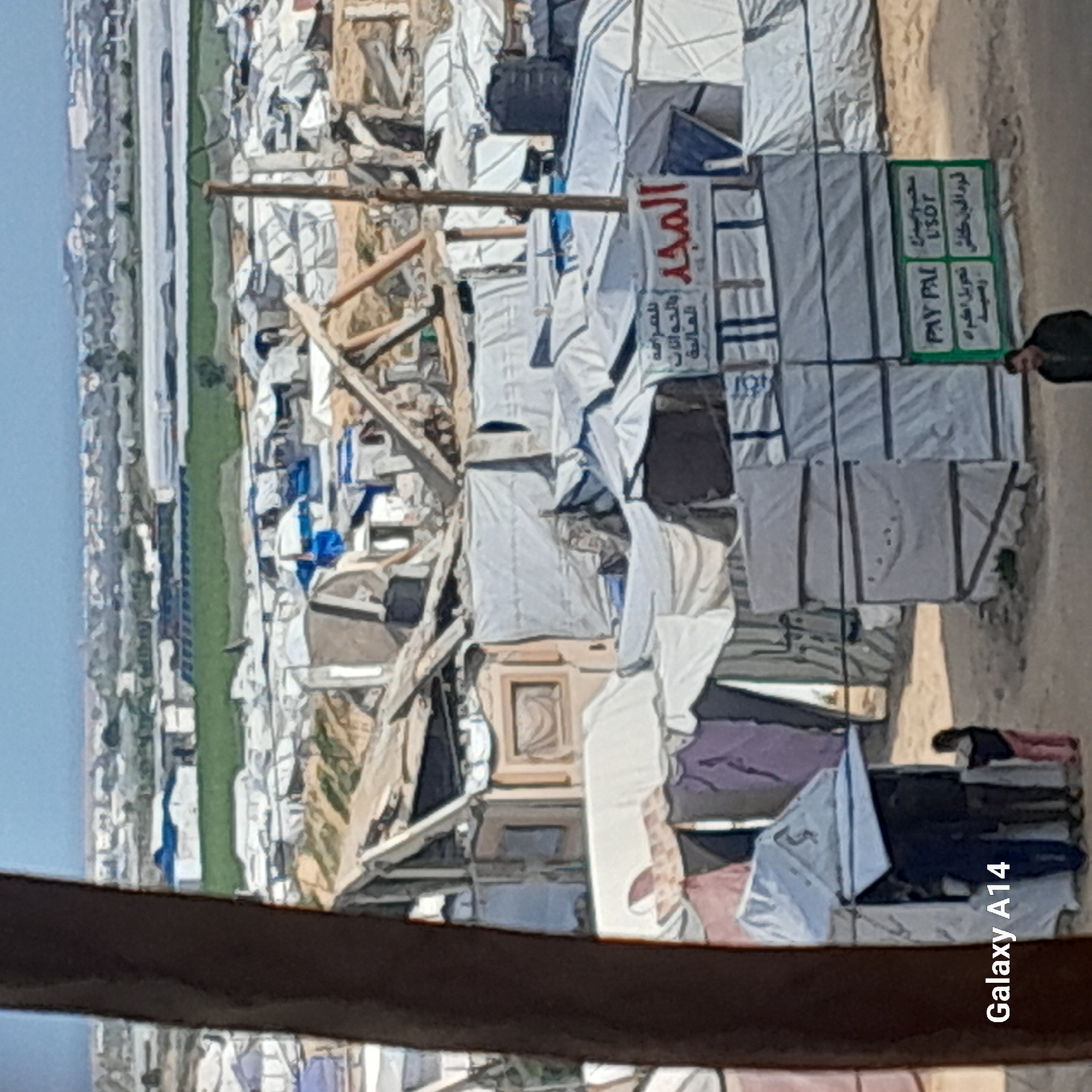 The image displays an elevated view of a densely populated informal settlement or camp in Khan Yunis, Palestinian Territory. The foreground consists of dirt ground, partially obscured on the left by a dark brown vertical pillar or beam.
The midground is dominated by a sprawling array of makeshift shelters constructed from white, gray, and blue tarpaulins or fabric sheets stretched over frames, some exhibiting exposed wooden structural elements, suggesting partial damage or unfinished construction. Numerous black cylindrical water tanks are visible on the roofs of various shelters. A prominent sign with a red background and white Arabic text reading "الْمَجْد" (Al-Majd) is affixed to a structure in the mid-right. Below this, a green and white sign displays "PAY PAL" and "USDT" alongside Arabic script, indicating money transfer or exchange services.
In the lower left, three individuals are present: two adults in dark clothing are positioned near a small, white tent-like structure with an open flap, and a child in pink pants and a dark top stands nearby. In the lower right, an adult male in a dark jacket is visible, facing towards the left.
The background features a distinct strip of green agricultural fields, beyond which lies a distant urban or semi-urban area under a hazy, light blue sky. The image quality is consistent with a mobile phone photograph, indicated by the "Galaxy A14" watermark in the lower left.