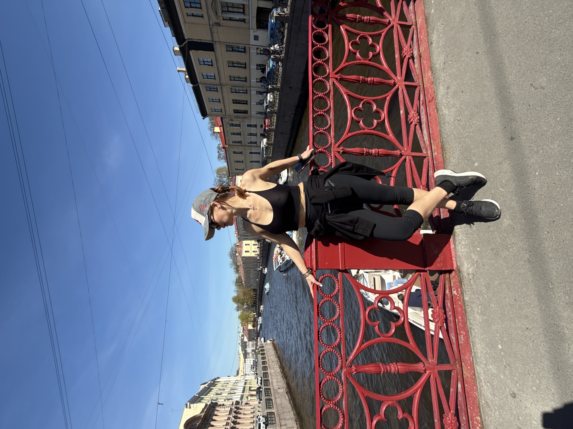 A rotated outdoor medium shot depicts a woman wearing a black tank top, black pants, black sneakers, and a gray hat, lying prone on a red bridge. The location is in Saint Petersburg, Russia, as indicated by the architecture. The bridge's railing features decorative cloverleaf patterns. Below the bridge is a canal with boats. Buildings line the canal, and a clear blue sky is visible above.