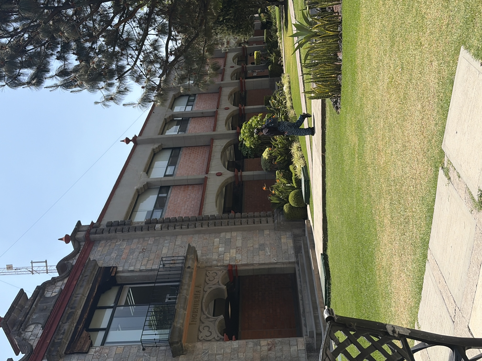 A daytime, eye-level shot shows a section of a three-story building and surrounding landscape in San Andrés Cholula, Mexico. The building features a mix of stone and brickwork, arched windows, and iron balconies. "INGENIERIA A" is etched above an arched doorway. To the building's right is a green lawn bordered by garden plants. A woman in a spotted outfit walks on the path next to the lawn. In the foreground, part of an iron railing is visible. A tree branch with pine needles is in the top-left corner. The sky is light blue. A crane is visible in the background.