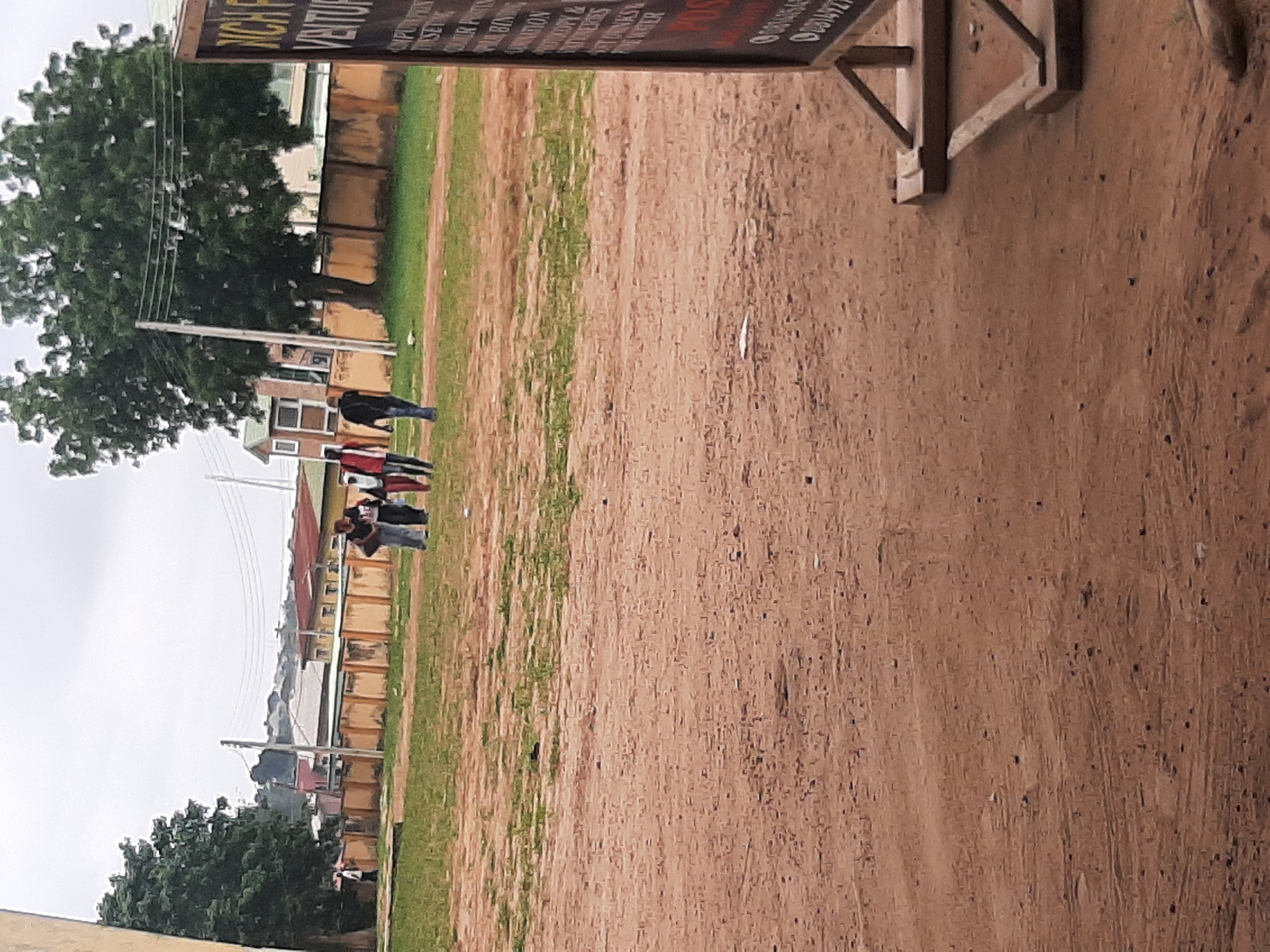 A group of four individuals, three females and one male, are walking across a dusty, open field in Ogbomosho, Nigeria. The females are wearing predominantly red and white outfits, while the male is dressed in black. In the background, a wall with a fence sits in front of several buildings. A large tree with lush green foliage stands to the left of the frame, with utility poles and wires visible behind it. A signpost is partially visible on the right side of the image, with some text on it. The sky is overcast and appears to be cloudy. The ground is mostly dry and sandy with patches of dry grass.