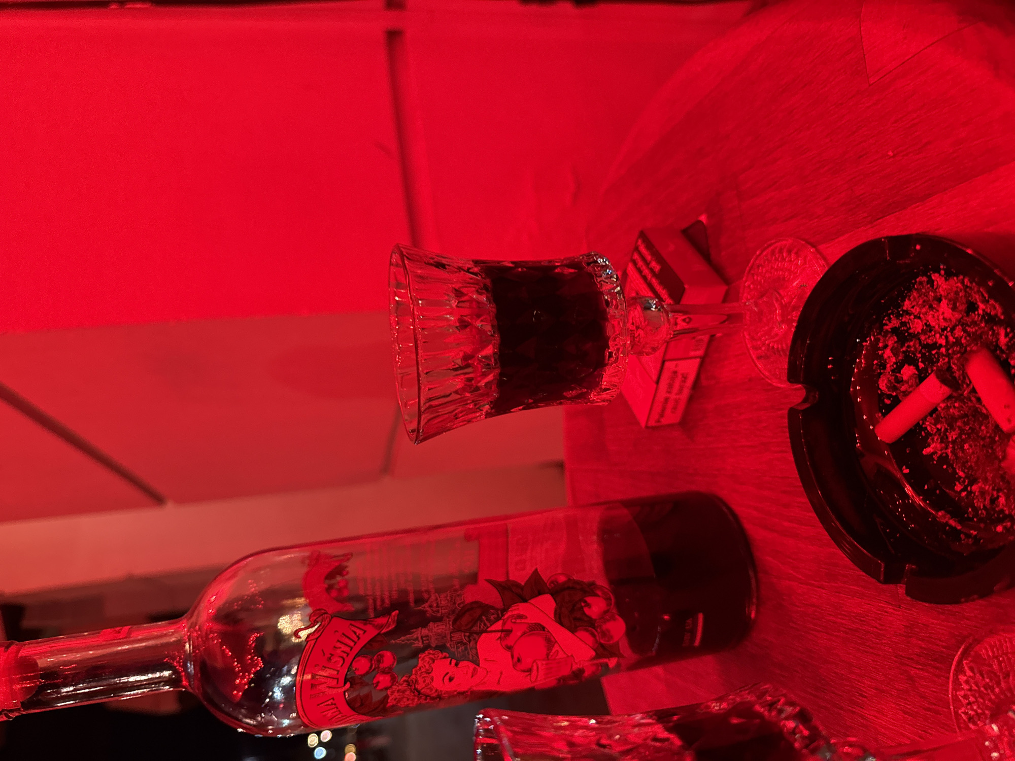 A bottle of "Maja Wisnia" liqueur, a glass filled with dark liquid, an ashtray with cigarette butts, and a pack of cigarettes are arranged on a wooden surface. The scene is illuminated by red lighting, casting a strong red hue over all the objects. The background appears to be a wall or building with a geometric design, also bathed in red light. The lighting suggests a nightlife setting, potentially a bar or club.