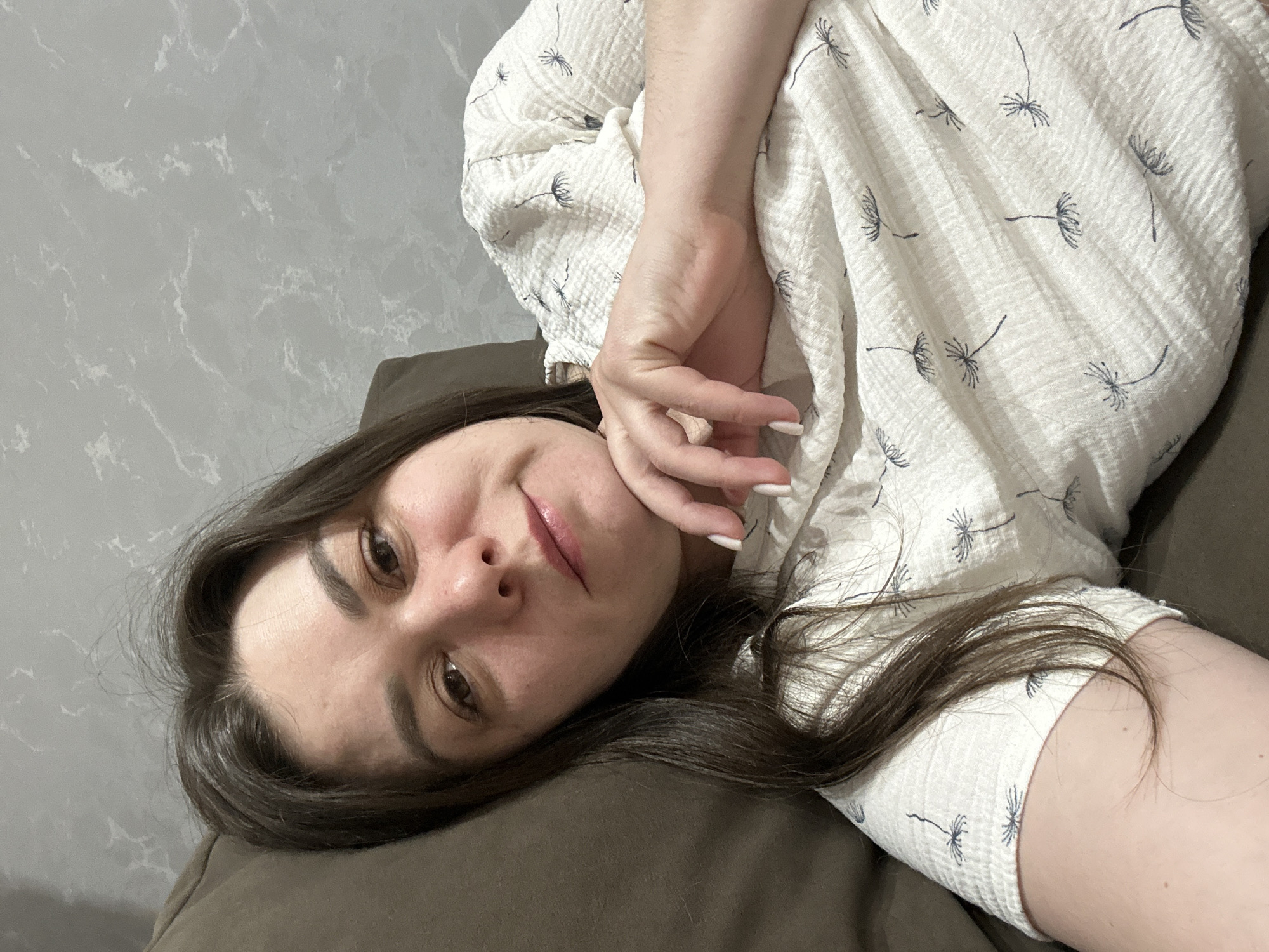 A woman is lying on a brown cushion in what appears to be an indoor setting. Her face is angled towards the camera, with her hand partially covering her cheek. She has dark hair and is wearing a white patterned blouse. The background features a gray wall with a mottled texture. There is no information to infer the location is in the city of Chaikovsky, Russia.