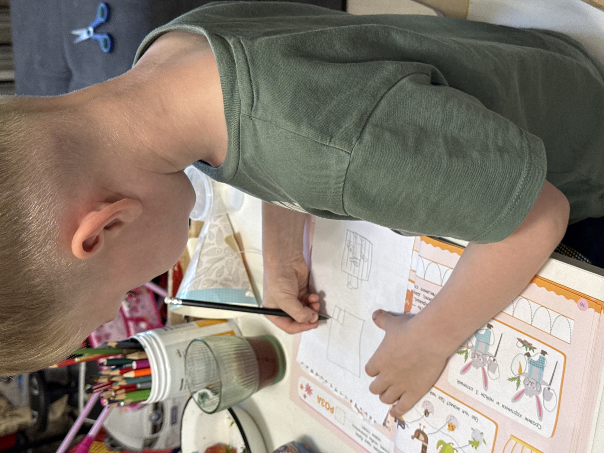 The image shows a close-up of a young boy with blonde hair, seen from the side and slightly from behind, as he is engaged in drawing on a pink and white activity book. He is wearing a green t-shirt. His hands are holding a black pencil and actively drawing on the paper. The activity book has illustrations of cartoon rabbits and text in Russian. In the background, there are colored pencils in a white container, a glass with a dark liquid, and other stationery items. A pair of blue scissors is visible on a dark surface in the far background. The scene appears to be a desk or table, suggesting an indoor setting, likely a home or classroom. There is no specific indication of the city of Chaikovsky, Russia, within the image itself.