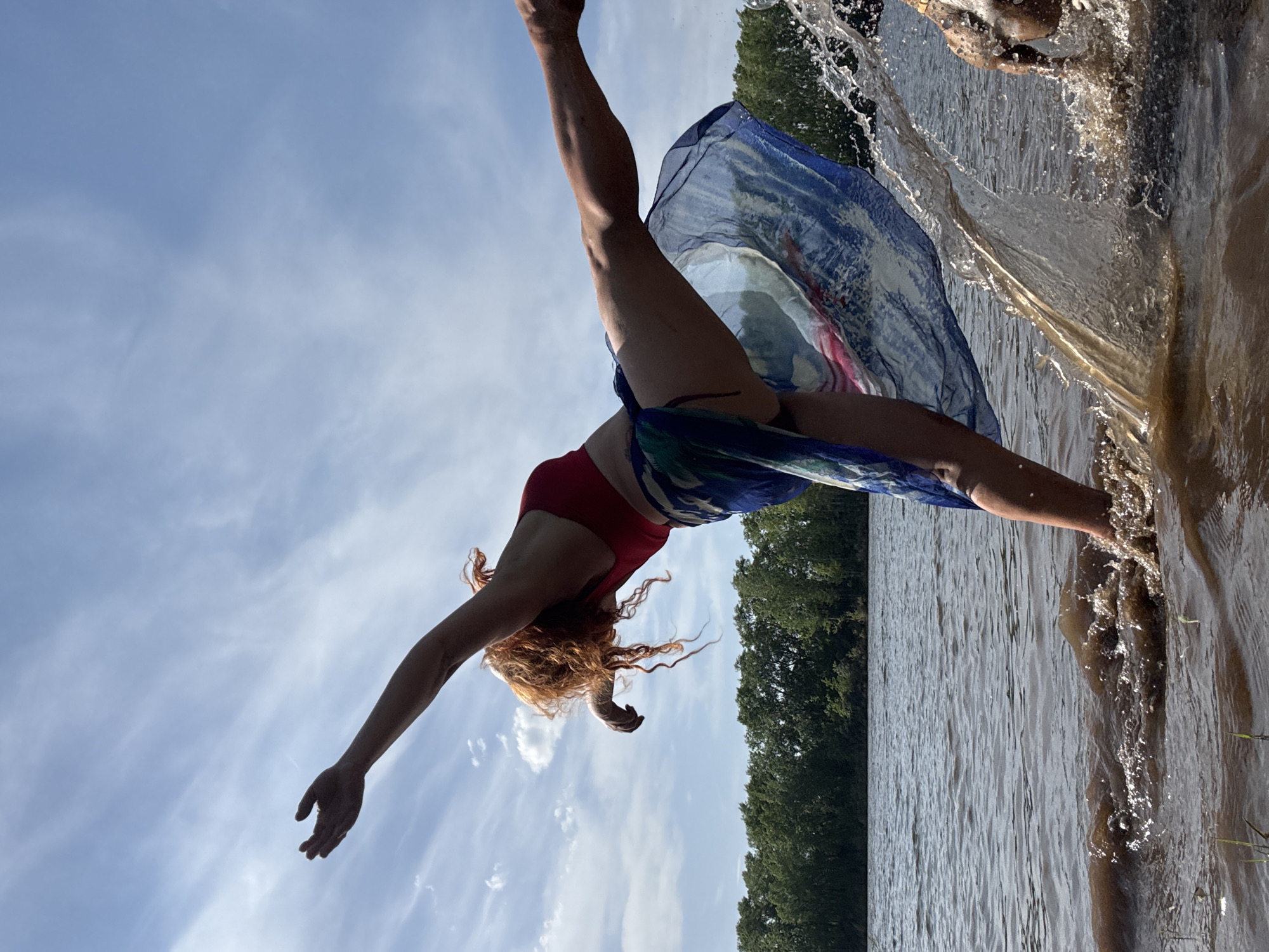 A fair-skinned female with red hair is diving headfirst into a body of water. She is wearing a red bathing suit and a flowing, blue and green scarf. Her right arm is extended toward the sky, and her legs are raised upward. Splashes of water are visible around her lower body, indicating the point of entry. The background features a line of green trees along the shoreline and a cloudy, blue sky. The location is in the city of Chaikovsky, Russia.
