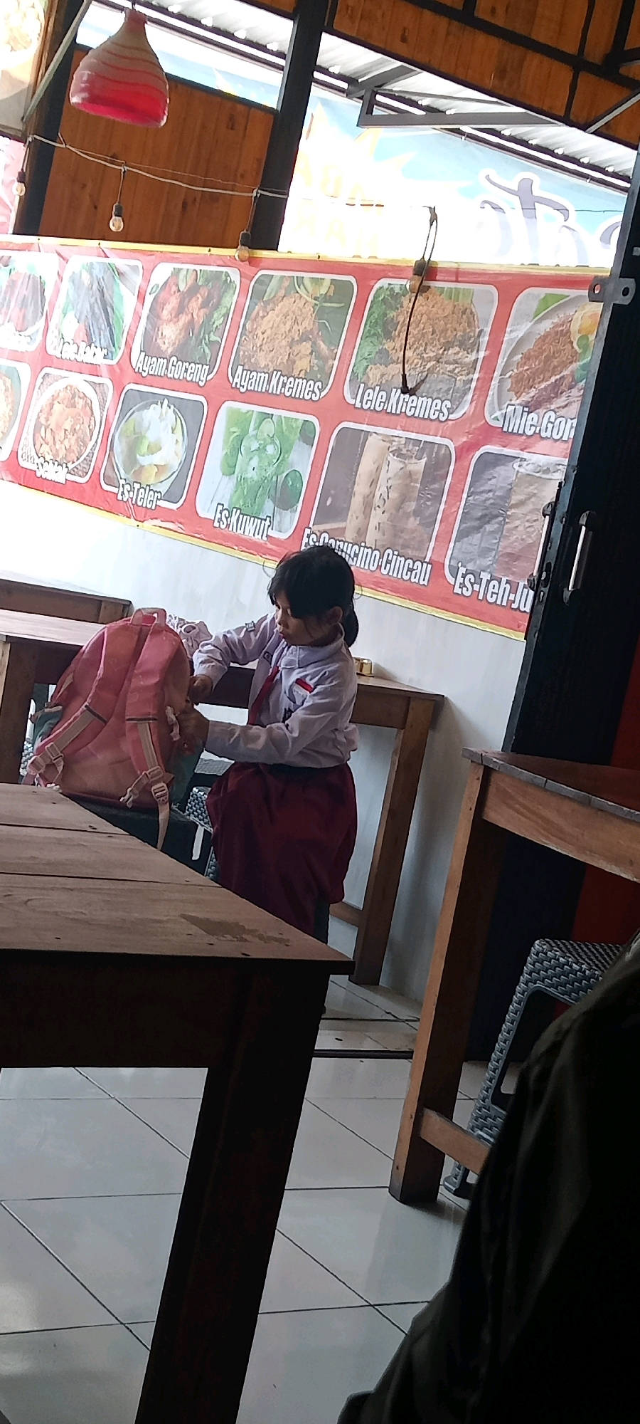 The image depicts an indoor setting, possibly a restaurant or food stall, with a young girl wearing a white and red uniform sitting at a wooden table. She has a pink backpack beside her and appears to be reaching into it. Behind her, a large banner displays various food items, including "Ayam Goreng" (fried chicken), "Ayam Kremes" (crumbed fried chicken), "Lele Kremes" (crumbed catfish), and "Mie Goreng" (fried noodles). The environment suggests a casual dining establishment, likely in Pondok, Indonesia, given the Indonesian text on the banner. The lighting is ambient, with a red decorative lamp visible in the background. The floor is tiled in a light color with a grid pattern.