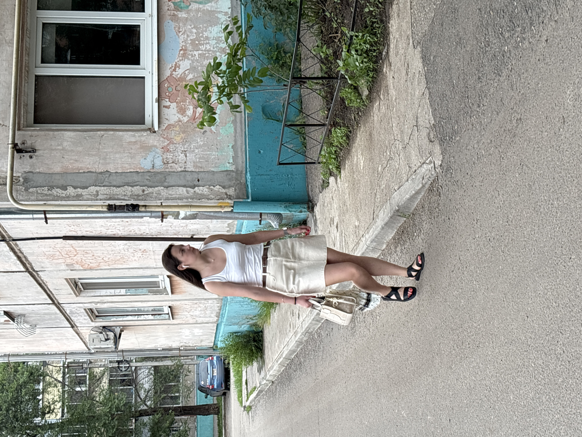 A woman stands on a paved surface next to a weathered, old building. She is wearing a white tank top and a beige skirt, with black sandals on her feet. She carries a light-colored handbag. The building behind her has peeling paint, showing multiple layers of color underneath. There are windows visible on the building's facade, and some pipes run along its exterior. Greenery, including small trees and bushes, is visible near the building and in the background. The ground is a mix of concrete and asphalt. A blue car is parked on the left side of the frame. The lighting suggests it is daytime. The image appears to be taken in an urban or suburban setting, possibly in Blagoveshchensk, Russia, given the context.
