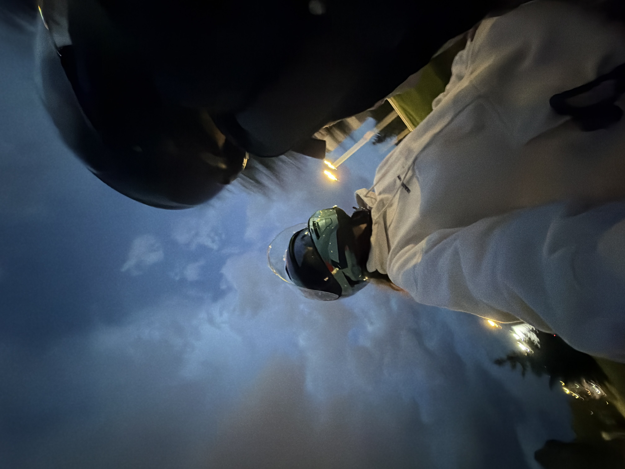 A low-angle, rotated image depicts two individuals against a dark, cloudy sky, likely taken at dusk or night in Sofia, Bulgaria. The perspective is from the ground looking upwards.
In the foreground, the top and side of a dark, glossy helmet are visible, belonging to one person whose body is mostly out of frame. Further back and to the right, a second individual is seen wearing a light-colored, possibly white or light grey, jacket and a distinctive green helmet with a clear visor. Their posture suggests they are either riding a motorcycle or scooter, or standing beside one, looking forward or slightly upwards.
The background features a dramatic, overcast sky with varying shades of dark blue and grey clouds. Several bright artificial light sources, possibly streetlights, are visible in the mid-ground and background, creating luminous flares and streaks of light against the dim environment. There is no visible text or identifiable architectural features beyond the general urban lighting. The scene conveys a sense of movement or being outdoors in the evening.