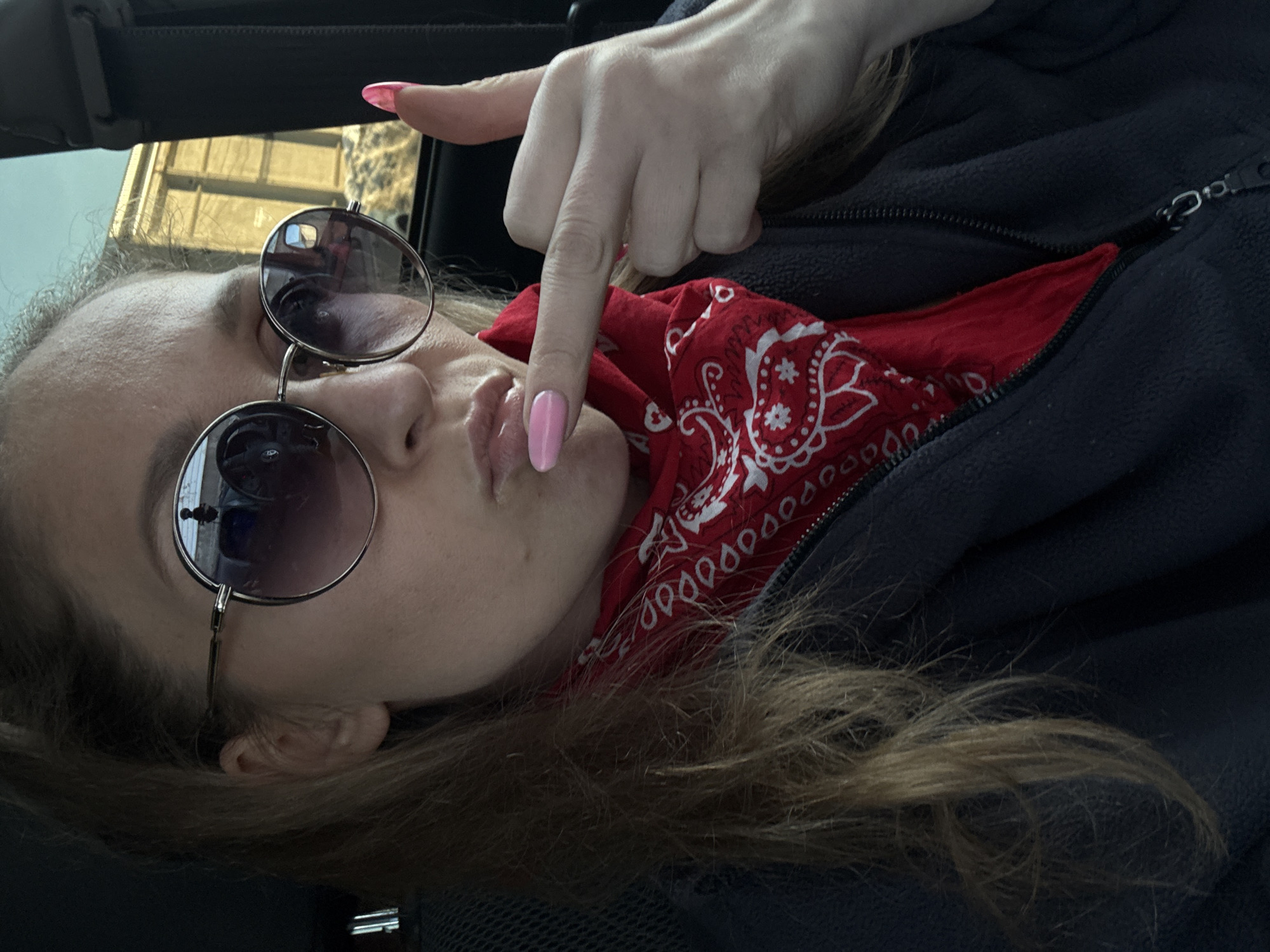 A person with fair skin, light brown hair, and pink nail polish is seated in what appears to be a vehicle, possibly a car. They are wearing round, dark-tinted sunglasses. The person is partially zipped up in a dark fleece jacket, with a red bandana visible at the neck. Their lips are pursed, and they are holding a finger near their mouth. The environment is dimly lit, suggesting indoor lighting or overcast conditions. The location is stated to be Chaikovsky, Russia, though there are no readily identifiable landmarks in the image to confirm this.