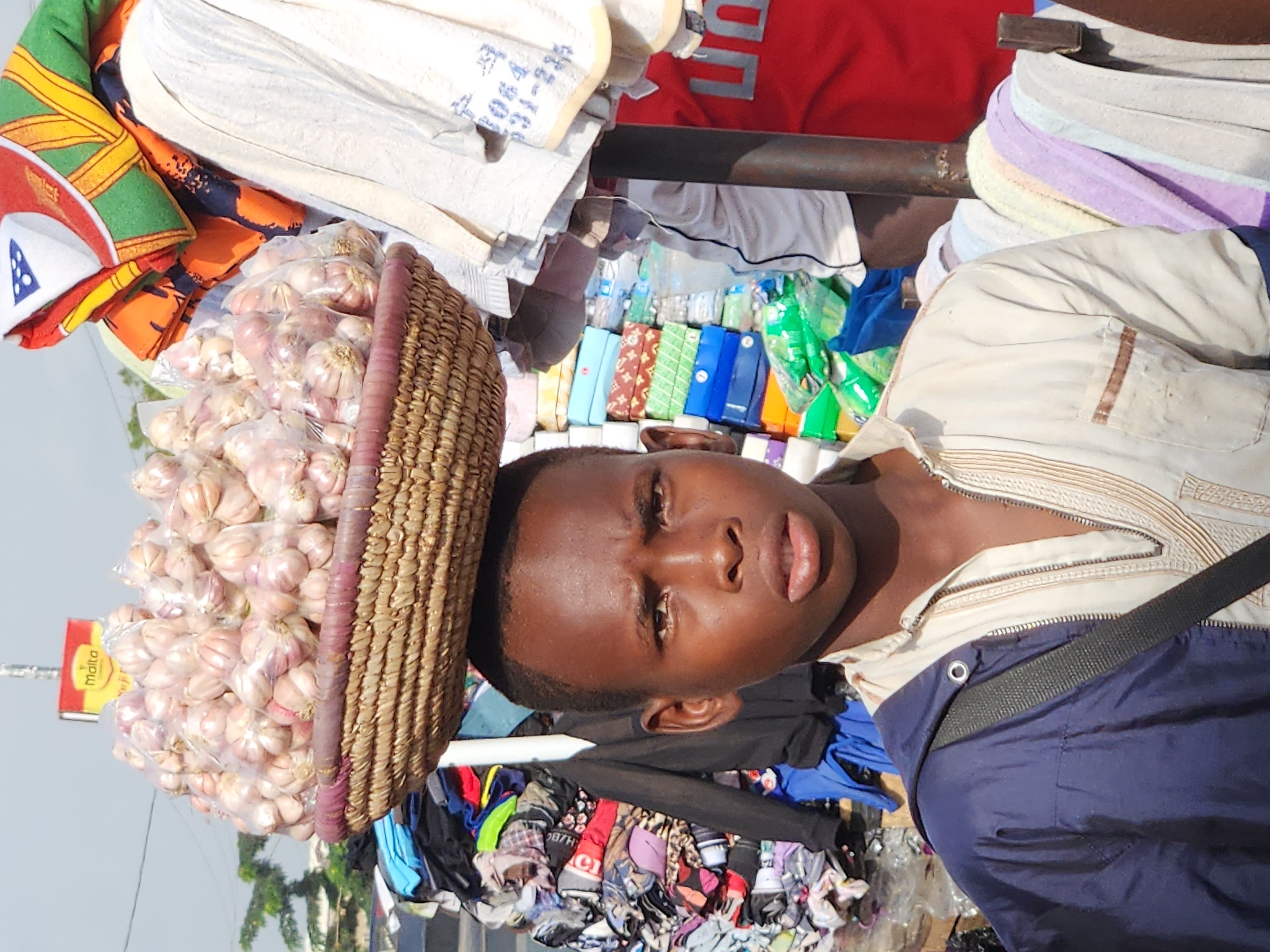 This is a vertical, eye-level, medium shot of a young Black person with dark skin, brown eyes, and dark hair. The person is wearing a light-colored, embroidered garment over a navy blue jacket with a black strap. Their head is tilted slightly to the right.
To the left of the person's head is a woven basket overflowing with garlic bulbs, which are individually bagged in clear plastic. The basket appears to be resting on something out of frame, and the person's head is positioned just below and to the right of it. In the background, there is a collection of colorful items, possibly clothing and other goods, stacked in a disorganized manner. Some of these items appear to be folded, while others are loose. A red object with white lettering is visible in the upper background, and a metal pole runs horizontally across the middle of the frame. There is a yellow sign with red text visible to the far left.
The lighting suggests it is daytime, possibly mid-morning or afternoon, with bright sunlight. The overall scene appears to be an outdoor market or street vendor setting in Sabon Tasha, Nigeria, indicated by the mention of the location context. The person's expression is neutral, and the overall mood is one of everyday commerce.