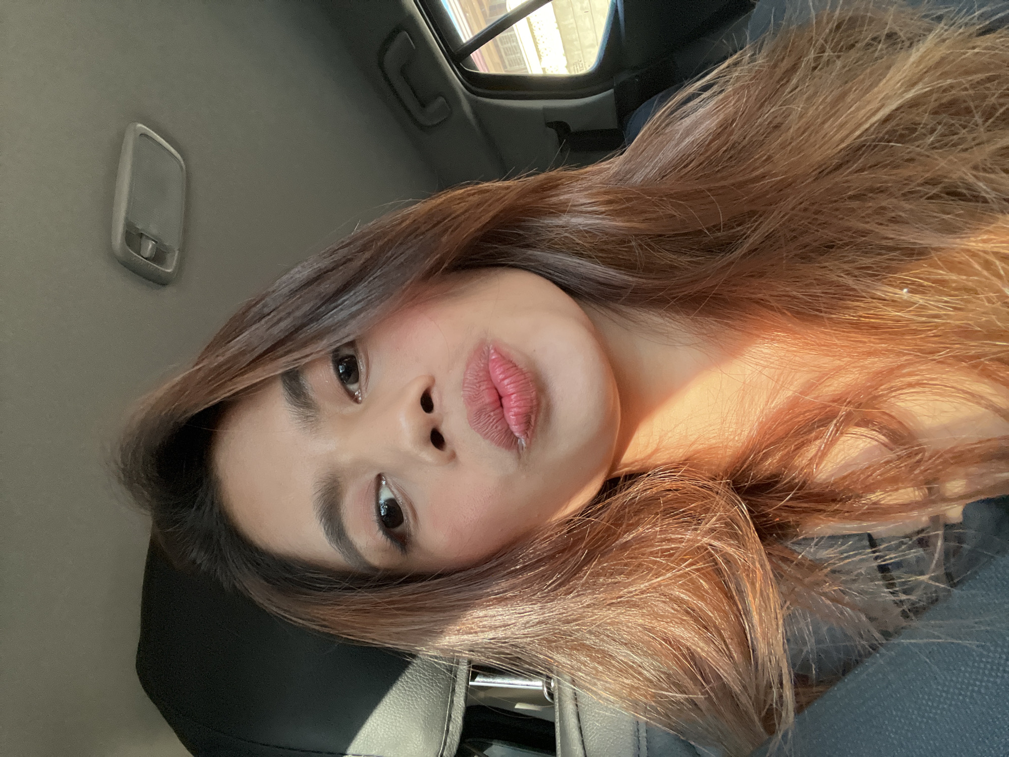 A young adult female is captured in a selfie-style photograph inside a vehicle in Davao City, Philippines. She is positioned looking directly at the camera with her lips pursed, an expression commonly known as a "duck face." Her long, brown hair with lighter auburn or golden-brown highlights cascades over her shoulders and chest, illuminated by strong natural sunlight entering from the left, casting a warm glow on her hair, right cheek, and neck. She wears a dark-colored top. The vehicle's interior features a black seat headrest to the left, a light-colored (grey or beige) headliner, and an overhead rectangular dome light with a toggle switch mechanism visible in the upper right. Through the side window on the right, a blurred exterior view shows elements of a building, including a light-colored wall, a window frame, and possibly a portion of a roofline, indicating an urban or suburban environment.