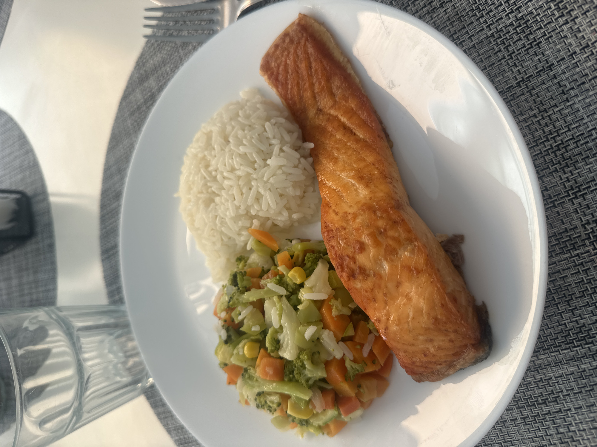 A high-angle shot captures a prepared meal served on a white, round plate, set on a dark gray woven placemat. The main subject is a large, golden-brown piece of cooked salmon, appearing crispy on top, which dominates a significant portion of the plate. Beside the salmon is a neat mound of white, long-grain rice. Below the rice and partially beneath the salmon, a colorful mix of steamed or sautéed vegetables is arranged, consisting predominantly of chopped green broccoli, diced orange carrots, and yellow corn kernels. Some white rice grains are visible among the vegetables.
In the upper left background, part of a silver fork is discernible, resting on the placemat. To the left of the plate, a clear glass tumbler with vertical ridges is partially visible. The scene is brightly lit by what appears to be natural daylight from the upper left, creating strong shadows on the plate and highlighting the textures of the food and placemat. The strong, directional lighting suggests it is daytime, possibly morning or afternoon. There are no people present, nor is any text visible in the image. The setting implies an indoor or covered dining area.