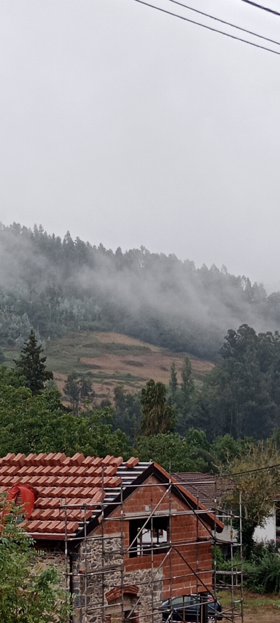 The image captures a cloudy, misty day in a rural setting, likely near Pravia, Spain. In the foreground, a house under construction is visible, with scaffolding surrounding it. The house has a stone base and brick walls, and its terracotta tiled roof is partially completed, with some tiles scattered. A red tarp is draped over a section of the roof. To the left of the house, lush green trees are abundant.
The background features a densely forested hillside shrouded in mist, creating a soft, diffused light. The trees on the hill appear to be a mix of evergreen and deciduous varieties. The mist lends a somewhat ethereal quality to the landscape. A patch of reddish-brown earth is visible on the slope of the hill, possibly indicating a cleared area or a different type of vegetation. The overall atmosphere is serene and damp, characteristic of a foggy, overcast day in a mountainous or hilly region. No people are visible in the image.