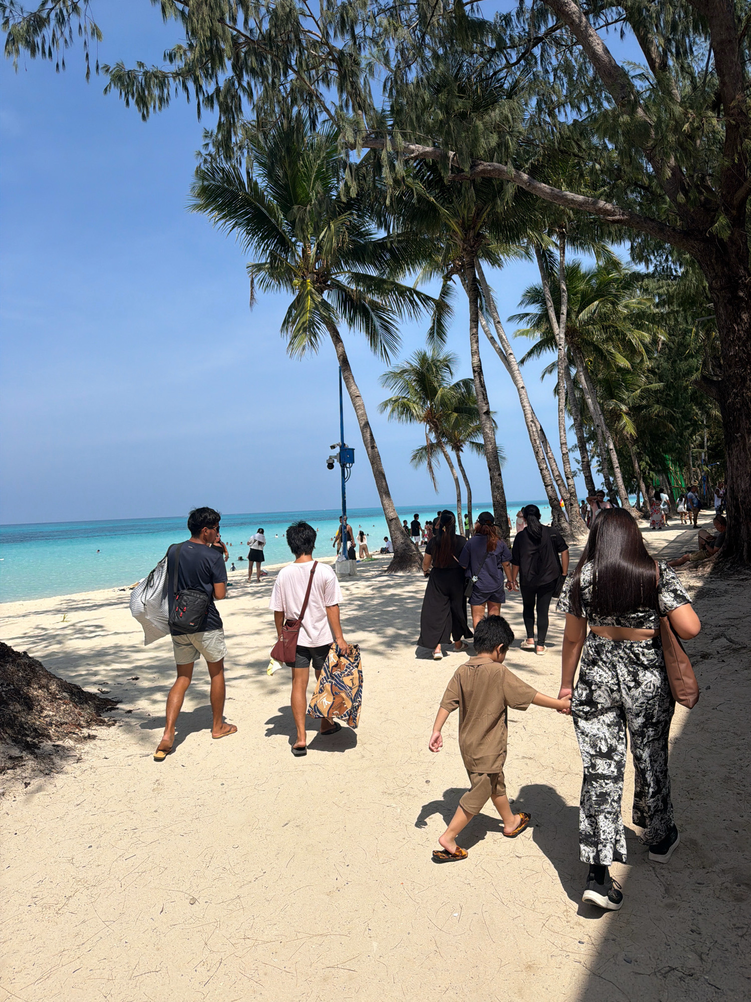 The image depicts a beach scene on a sunny day in Malay, Philippines. The foreground features a sandy path with scattered palm fronds and shadows cast by trees lining the right side of the frame. Several people are walking along the path, some from behind. Two young men, one with a dark t-shirt and shorts carrying a large striped bag, and another in a light pink t-shirt and shorts carrying a patterned bag, are walking away from the camera. Behind them, a group of women in dark attire are also walking. Further back, a young boy in a brown outfit is holding hands with a woman in a patterned two-piece outfit, and they are also walking away from the camera. The background reveals a pristine beach with turquoise water and several individuals swimming or relaxing. Palm trees are abundant, casting dappled shadows on the sand. The sky is a clear, bright blue.