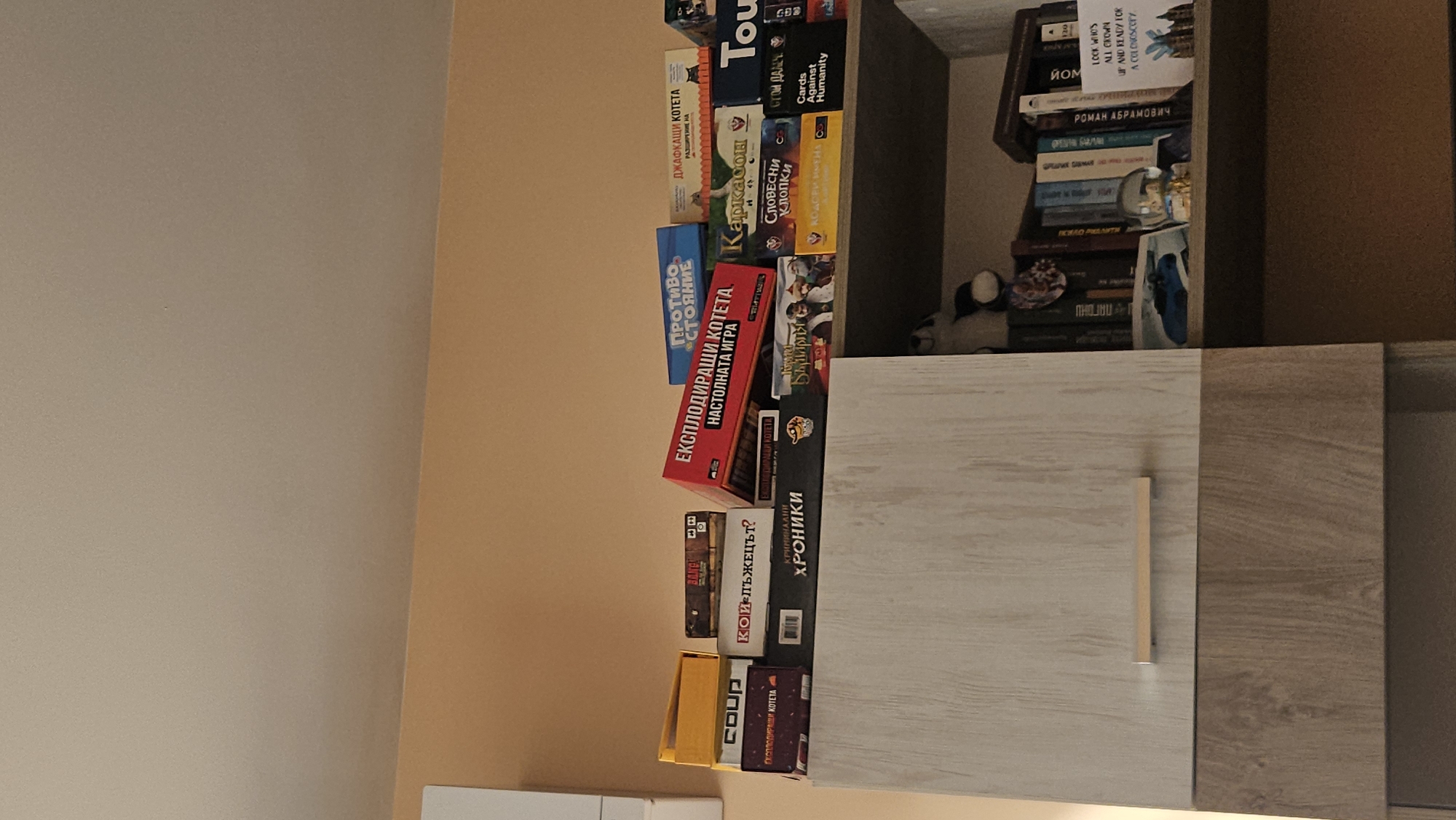 A tilted, close-up shot shows a cabinet filled with board games and books. The games include "Exploding Kittens," "Cards Against Humanity," and others with Cyrillic titles, such as "Джафкащи котета" and "Словесни Клопки." Titles on the books include "Роман Абрамович." A plush toy is partially visible among the books. The cabinet has light and dark wood-grain doors. The scene takes place indoors, likely in a home. The image is likely taken in Bulgaria based on the Cyrillic game titles, potentially in Plovdiv given the prevalence of Bulgarian language games.