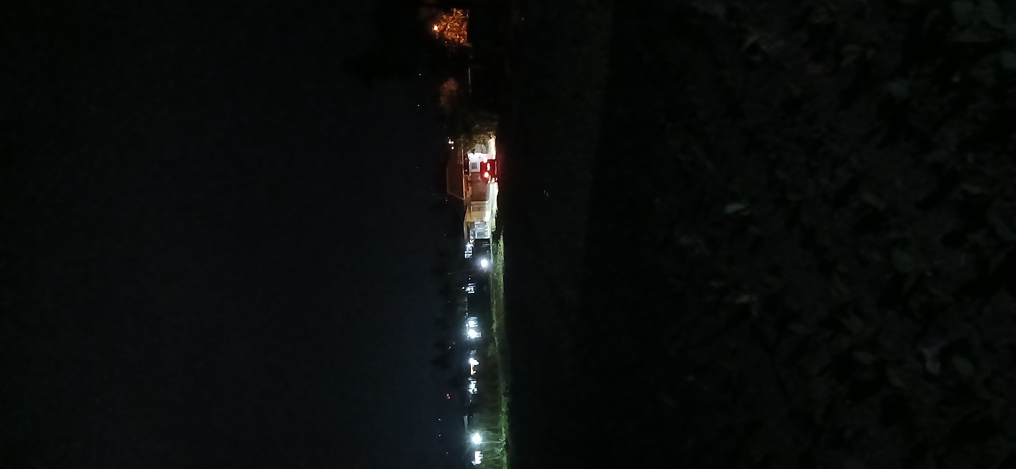 The image depicts a dimly lit outdoor scene at night, likely in Sekarputih, Indonesia. The foreground is dark and appears to be an open field with some vegetation. In the middle ground, a row of buildings is visible, illuminated by artificial lights. A red car is parked in front of one of these buildings, with its taillights illuminated. The background is a dark, starless sky. The overall impression is one of a rural or semi-rural area at night.