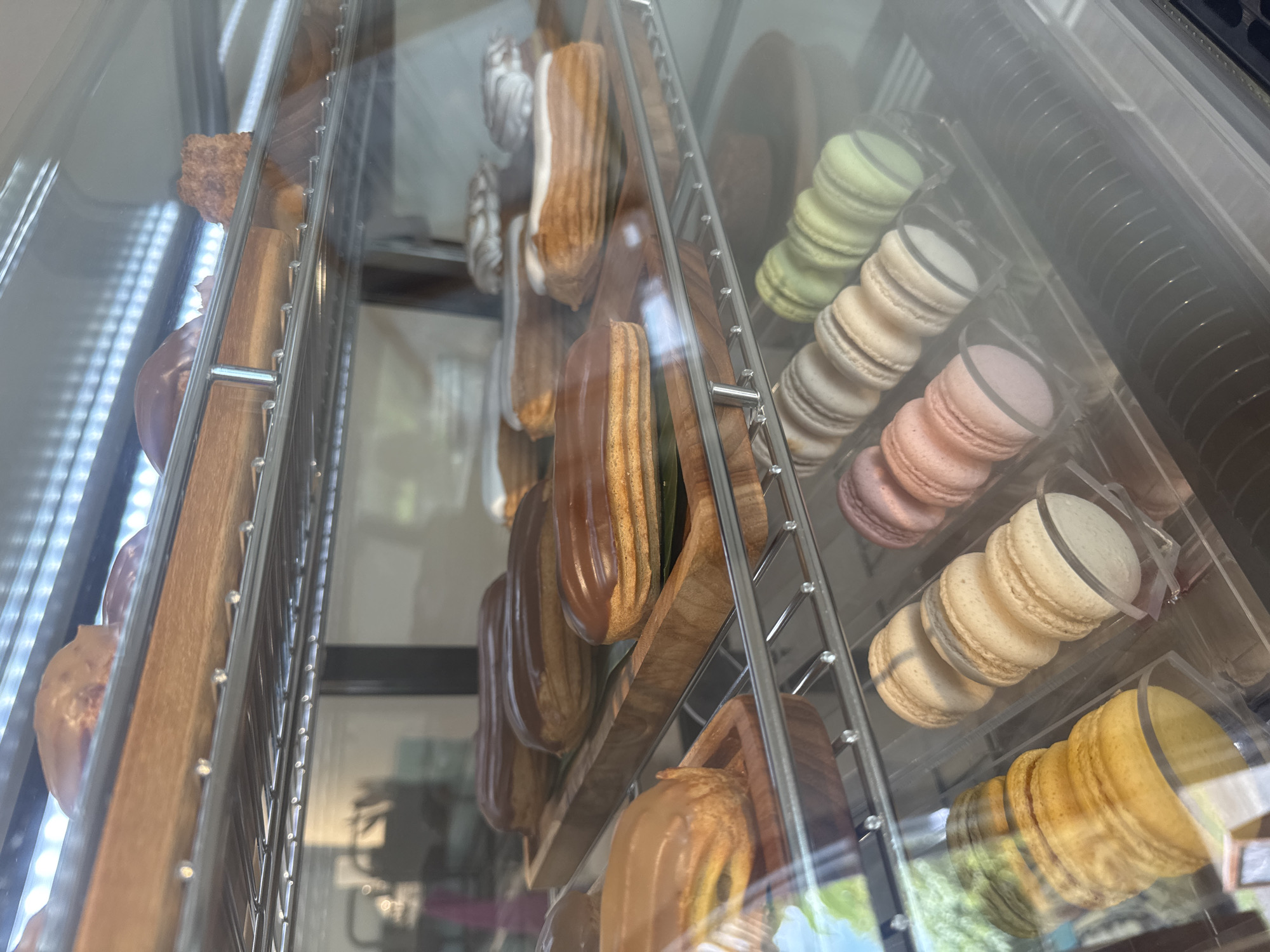 The image depicts a refrigerated display case filled with various baked goods, likely from a bakery or cafe. In the foreground, on the bottom shelf, are rows of macarons in different colors: yellow, beige, pink, and light green. Behind them, on a higher shelf, are eclairs with chocolate and white icing. Further up, another shelf holds more eclairs with chocolate icing. The display case is illuminated from above by fluorescent lights. The background is blurred but suggests an indoor setting with some daylight filtering through. There are no people visible in the image. The scene is clean and organized, suggesting a commercial establishment. There is no information in the image to indicate its specific location in Abang, Indonesia.