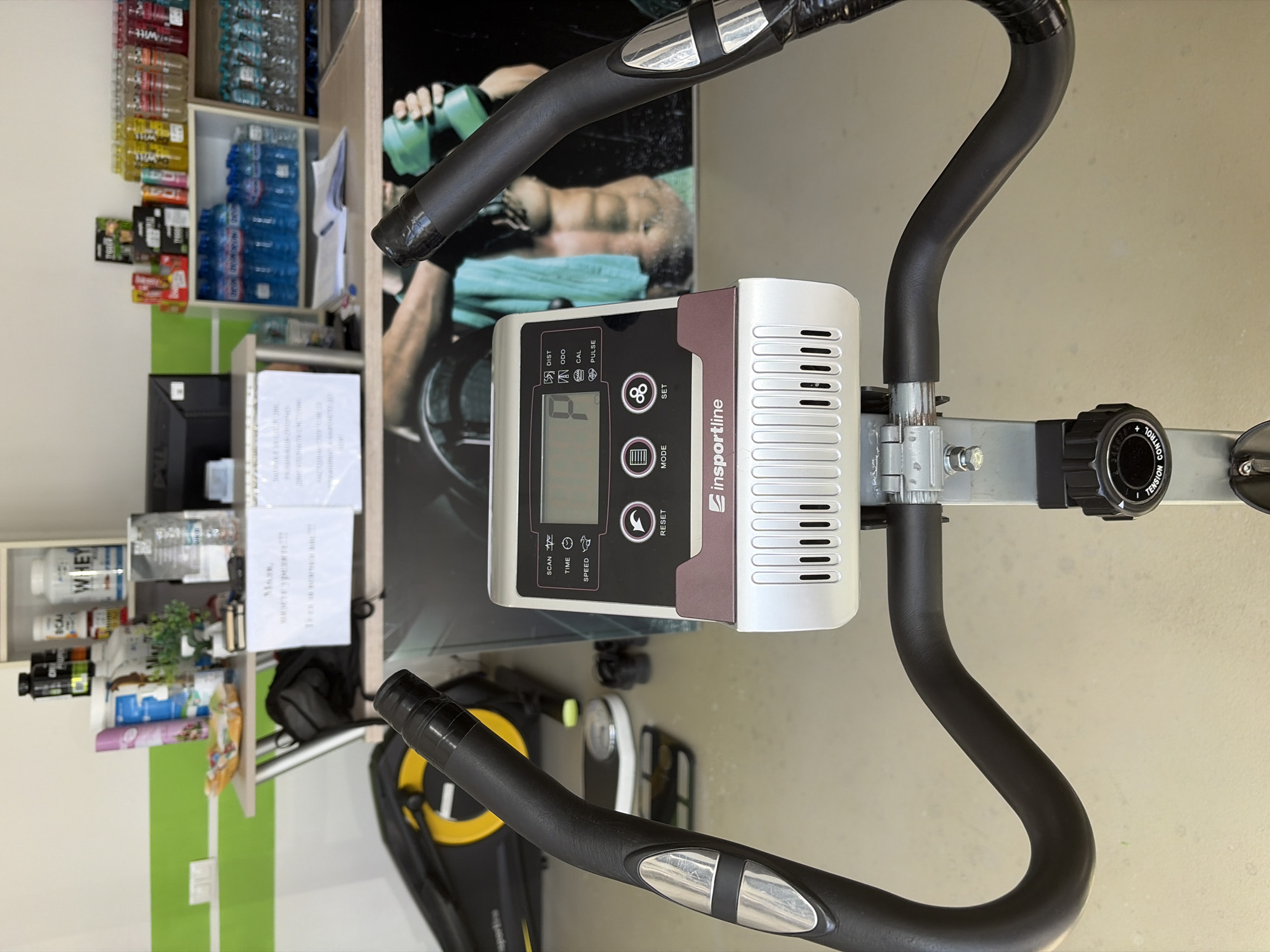 This is an indoor shot featuring the display and handlebars of a stationary exercise bike, possibly located in a gym or home exercise area. The display reads a value of '0' with labels for 'SCAN', 'TIME', 'SPEED', 'DIST', 'ODO', 'CAL', 'PULSE', and has buttons for 'MODE', 'RESET', and 'SET'. The brand name "insportline" is visible on the display unit. The handlebars are black with silver accents. A tension control knob is situated near the handlebars.
In the background, there is a shelf stocked with water bottles, various supplements and health products, and some paperwork. A poster of a muscular person is also visible. A black bag can be seen on a lower shelf. While the image was taken in Bulgaria is mentioned, there is no indication about the location in the city of Shumen in the image.