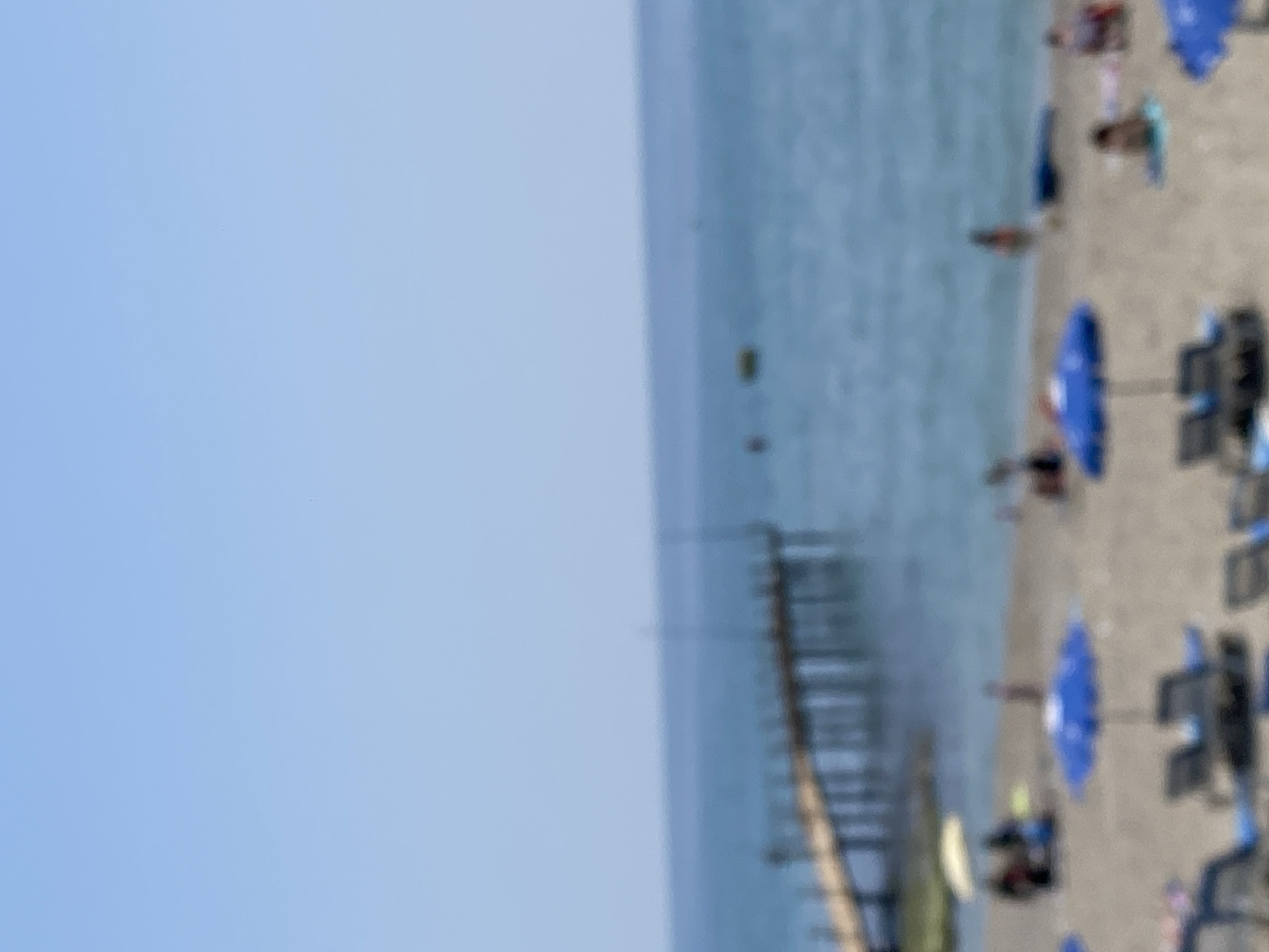 The image depicts a beach scene in Alanya, Turkey. The beach is populated with people, blue umbrellas, and lounge chairs. A pier extends into the sea. The sky is clear and blue.