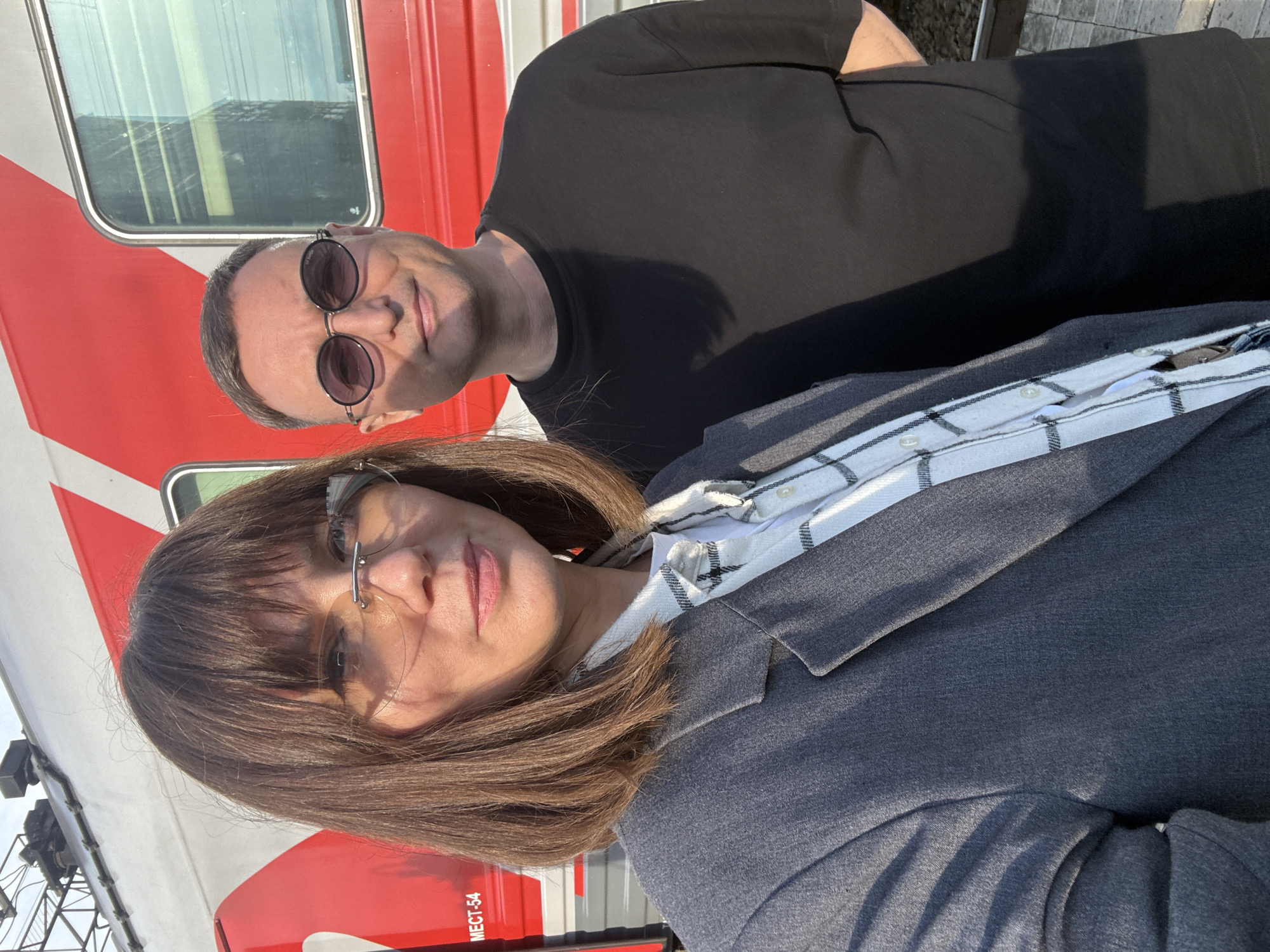 Two individuals, a woman and a man, are pictured outdoors, standing in front of a red and white train. The woman, on the left, wears a grey blazer over a white and black checkered shirt. She has medium-length brown hair and is wearing rimless glasses. The man, on the right, is wearing a black t-shirt and round, dark sunglasses. The train appears to be at a station platform, with visible tracks and ballast beneath their feet. The background suggests an urban or industrial setting, possibly related to railway operations in Izhevsk, Russia. The lighting indicates it is daytime, with sunlight casting shadows.