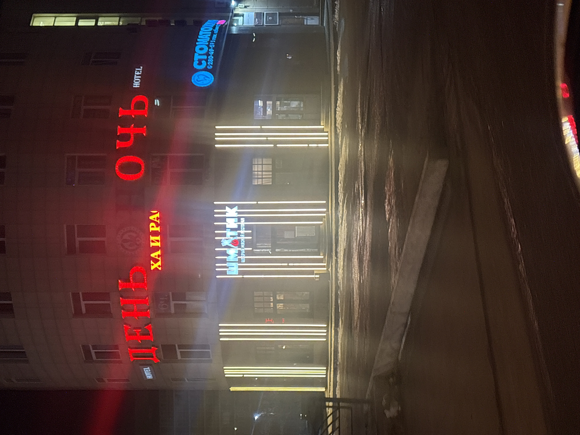 The image displays a nighttime street scene in Krasnoyarsk, Russia, focusing on the illuminated facade of a multi-story commercial building. The ground is wet and reflective, suggesting recent rain or snow, and amplifying the glow of the lights.
Prominently featured is a large vertical red neon sign on the building. On the left, it spells out "ДЕНЬ" (DAY), and on the right, "НОЧЬ" (NIGHT). Below "НОЧЬ", the word "HOTEL" is visible in red. Beneath "ДЕНЬ", smaller red text reads "ХАЙРА" (KHAIRA).
Higher on the building, a blue circular sign with a tooth logo clearly advertises "СТОМАТОЛОГ" (DENTIST), along with a phone number "280-49-59" and "3 этаж, кабинет" (3rd floor, office).
On the ground level, vertical strips of bright yellow lights frame an entrance. Above these lights, a sign identifies "ШМАТИК" as a "ЩЕДРЫЙ МЯСНЫЙ РЕСТОРАН" (GENEROUS MEAT RESTAURANT). Numerous windows are visible across the building's floors.
No people or vehicles are present, making the building and its vibrant neon signs the central subjects of this urban night view. The scene is static, highlighting the commercial activity through its illuminated signage.