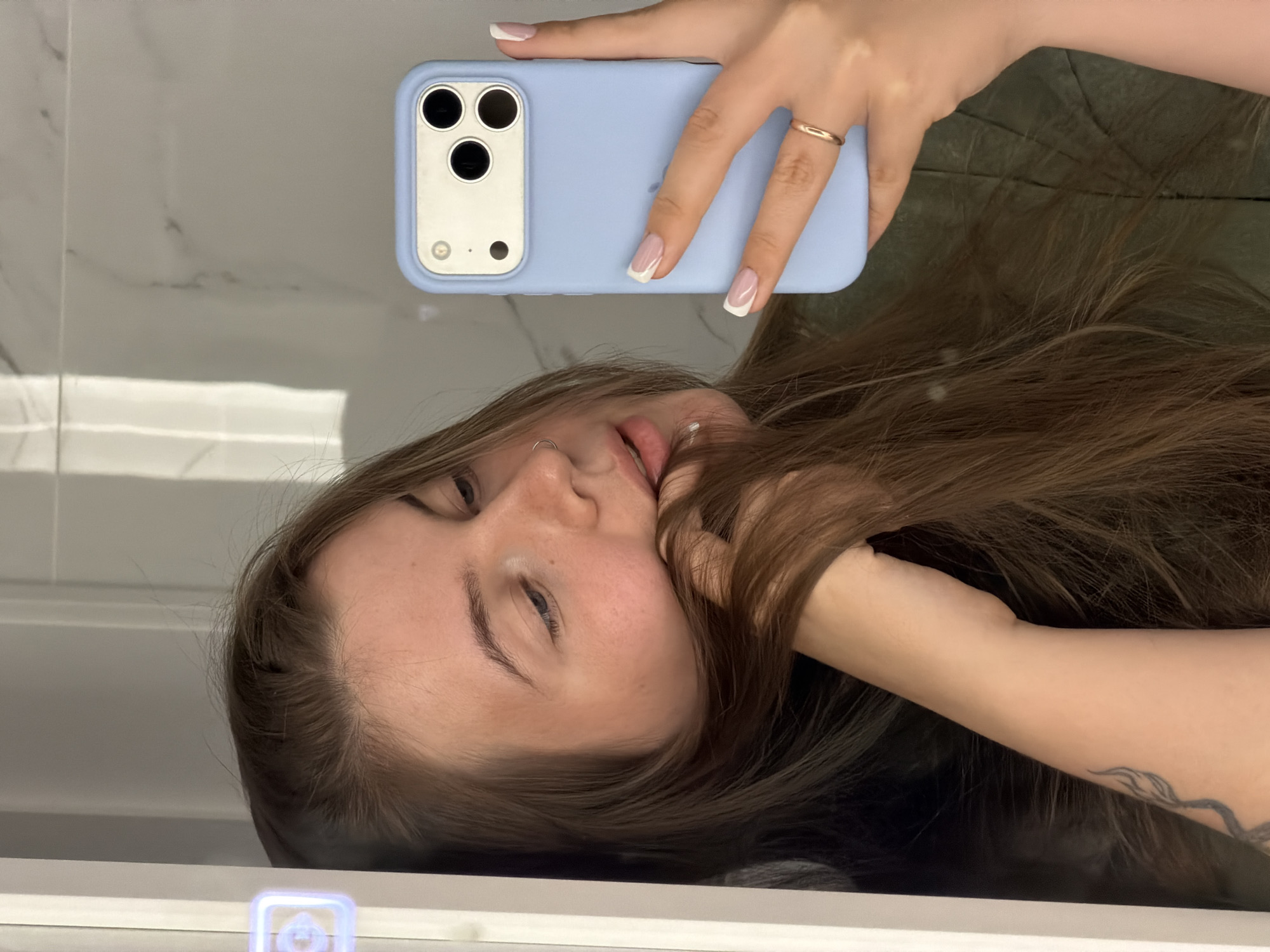 A woman is taking a selfie in a bathroom with a blue iPhone. The phone is in her right hand, and her fingers are visible. She is wearing a gold ring on her finger, and her nails are painted with a white tip. The woman's face is visible in the reflection of the mirror. She has long brown hair and a nose piercing. A tattoo is visible on her arm. The bathroom has white marble tiles and a white vanity. There are no indications of the image being taken in the city of Chaikovsky, Russia.