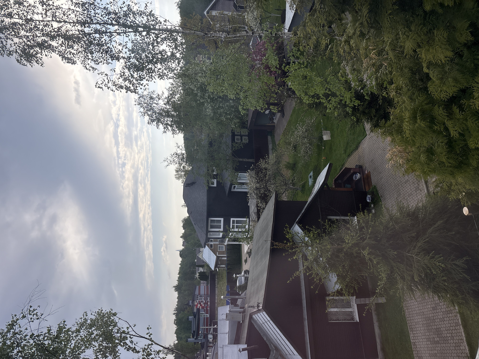 The image presents an elevated, slightly rotated view over a neighborhood consisting of residential buildings, likely located within Saint Petersburg, Russia. Structures are predominantly of a dark brown or black color, with white window frames. Several structures showcase dark roofs. The landscape is lush with trees and grass. A paved, curved walkway is also visible. The sky indicates a moderately cloudy day. No human presence is directly observable in the frame.