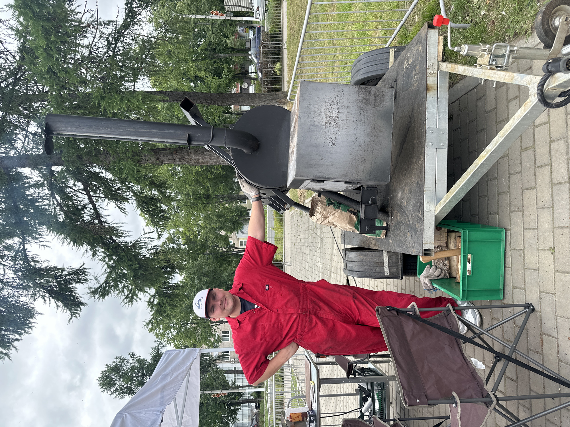 A man in a red jumpsuit and a white baseball cap stands next to a large, dark smoker on a trailer. He has his left hand on his hip and his right arm is bent, with his hand resting on the smoker. The smoker is black and has a tall chimney. The trailer has two wheels and a metal frame. In the background, there are trees, buildings, and a fence. There is a white tent to the left of the man, and a folding chair in the foreground. A green crate filled with firewood and tools is next to the trailer. The scene appears to be outdoors, possibly at a festival or event. The location is the city of Chaikovsky, Russia, specifically near the embankment of the Kama River.