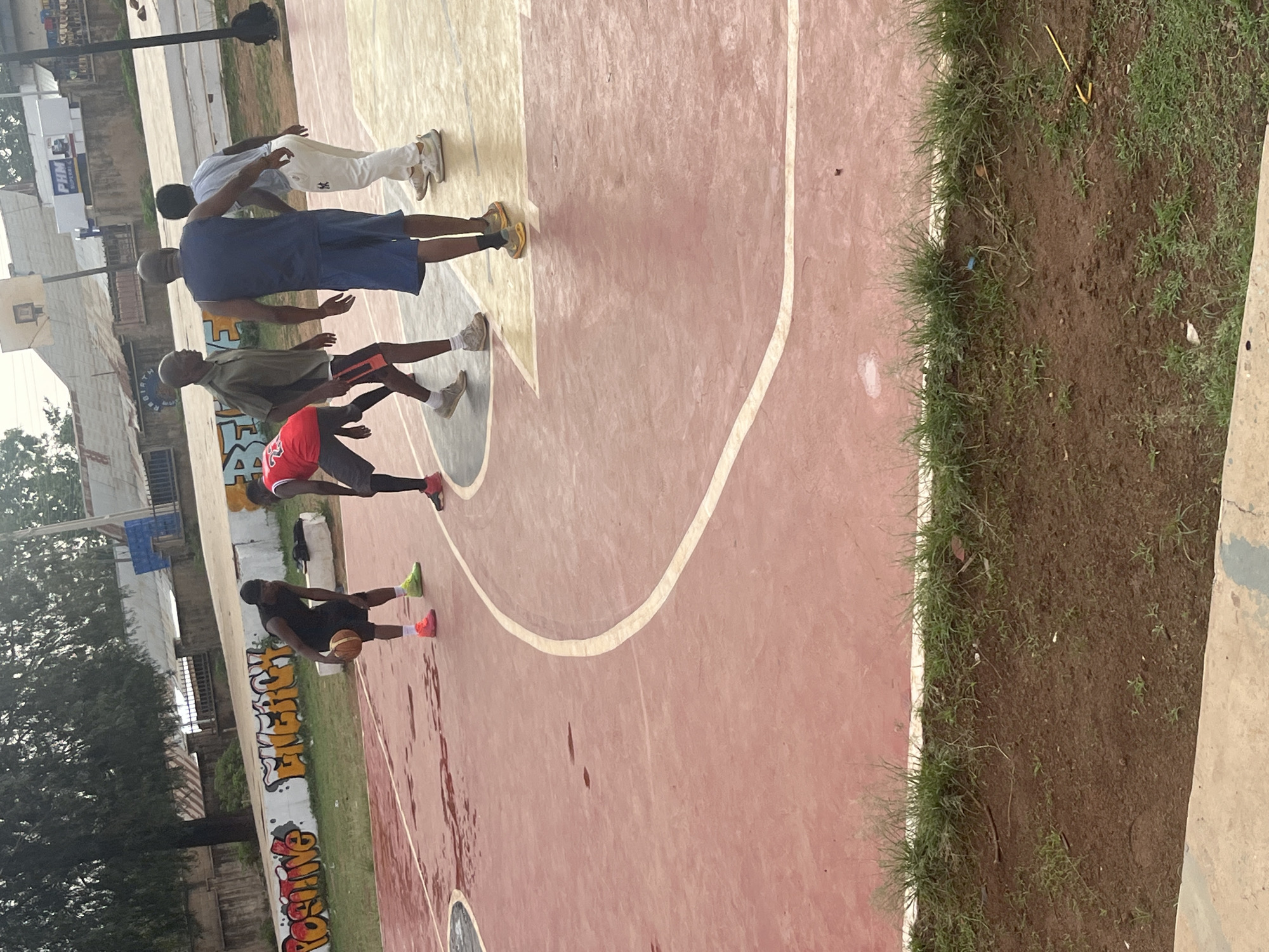 A medium shot depicts five people playing basketball on an outdoor court. The court is painted with a red and yellow color scheme, and a white line marks the perimeter. A person in black shorts and a black shirt dribbles the ball. A person in a red shirt stands nearby. Three additional people are positioned on the court, including one in a blue shirt. Graffiti is visible along the edge of the court, near the grassy area. The location is an urban setting in Gura Top, Nigeria. Buildings and trees are visible in the background.