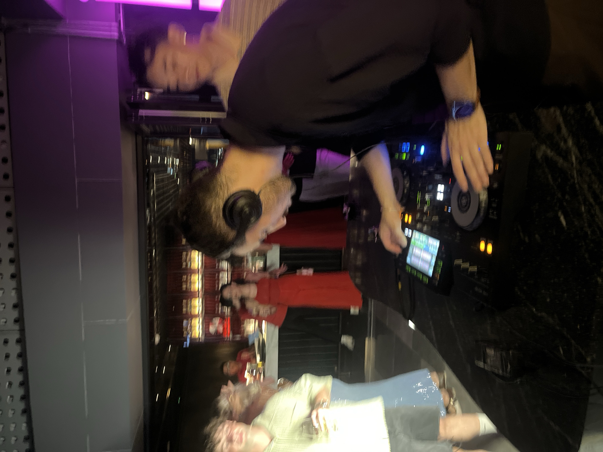 The image shows a group of people in what appears to be a dimly lit indoor venue, possibly a nightclub or bar, with purple lighting visible in the background. A person wearing headphones and a black t-shirt is standing behind a DJ setup, with their hands on the controls, suggesting they are the DJ. They are wearing a watch on their left wrist. In the background, other people are visible, some in red clothing. The scene suggests a social gathering or event, possibly a party. The orientation of the image is rotated, making it appear upside down. There are no discernible text elements in the image.