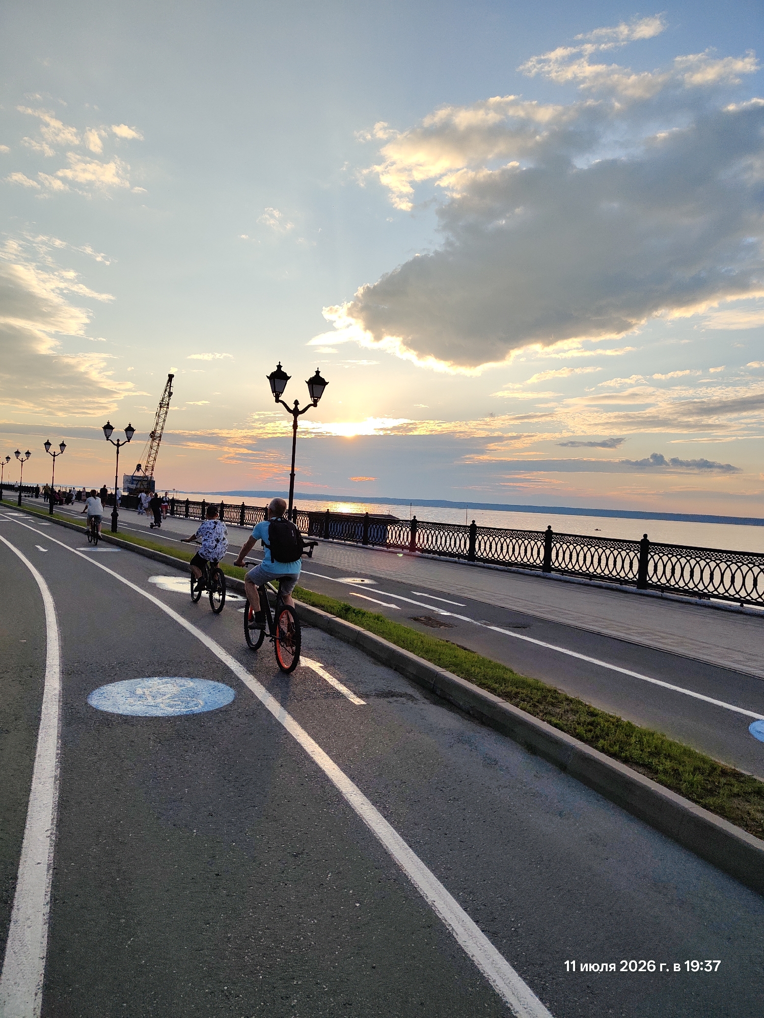 The image captures a sunset scene at a waterfront promenade. In the foreground, a paved path with white lane markings is visible, featuring a blue circular symbol indicating a bike path. Two individuals are riding bicycles on this path. The person in the foreground is wearing a blue t-shirt and shorts, with a black backpack, and is riding a bicycle with orange accents on the wheels. Behind them, another person is riding a bicycle. The path curves to the left, with more cyclists and pedestrians further in the distance. Along the right side of the path is a decorative metal fence separating it from a body of water. Beyond the water, a distant shoreline is visible, and the sky is filled with a dramatic sunset with illuminated clouds. In the background, a large crane is visible, suggesting ongoing construction or industrial activity. Several streetlights line the promenade. The timestamp "11 июля 2026 г. в 19:37" is superimposed on the lower right corner of the image.