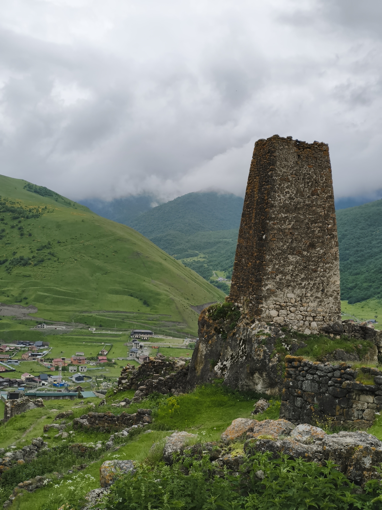 The image captures a view of a medieval stone tower in Verkhny Fiagdon, Russia, overlooking a village nestled in a lush green valley. The tower, constructed from rough-hewn stones with patches of moss and lichen, stands prominently on a rocky outcrop in the foreground. Its top is weathered and partially broken, suggesting age and exposure to the elements. Surrounding the tower are remnants of stone walls and scattered rocks, indicative of an ancient structure.
In the mid-ground, the village of Verkhny Fiagdon unfolds. Houses with red and brown roofs are clustered together, interspersed with green spaces and roads. The village is situated on a relatively flat area at the base of steep, verdant mountains.
The background is dominated by rolling green hills and mountains, shrouded in a soft, overcast sky with patches of mist clinging to the higher elevations. The landscape is characterized by its natural beauty and the harmonious coexistence of human settlement with the rugged mountainous terrain. The overall impression is one of historical significance and serene natural beauty.