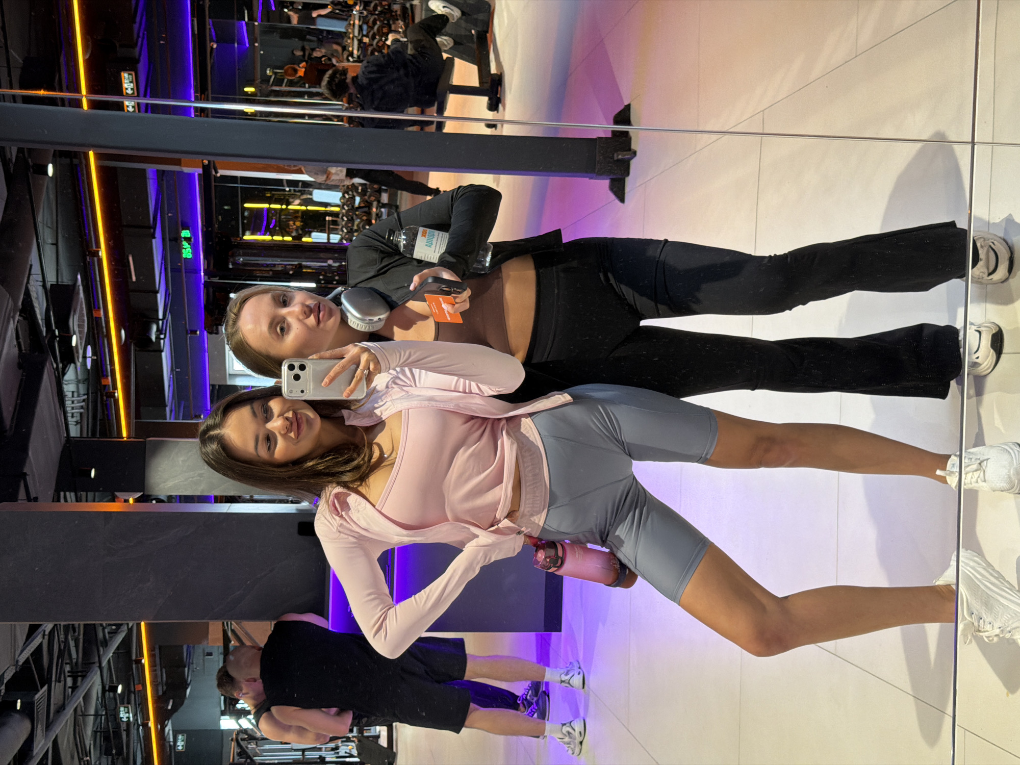 Two young women are posing for a selfie in what appears to be a gym in Krasnoyarsk, Russia. The gym equipment is visible in the background. The woman closer to the camera has long brown hair and is wearing a light pink jacket, blue shorts, and white shoes. She is holding a white smartphone to take the picture. The other woman is wearing black pants, a black top, and headphones around her neck. She is holding a bottle of water and a small orange card. Both women are smiling. The gym has purple and orange lighting. Other people are working out in the background. The floor is white and reflective, showing reflections of people and objects.
