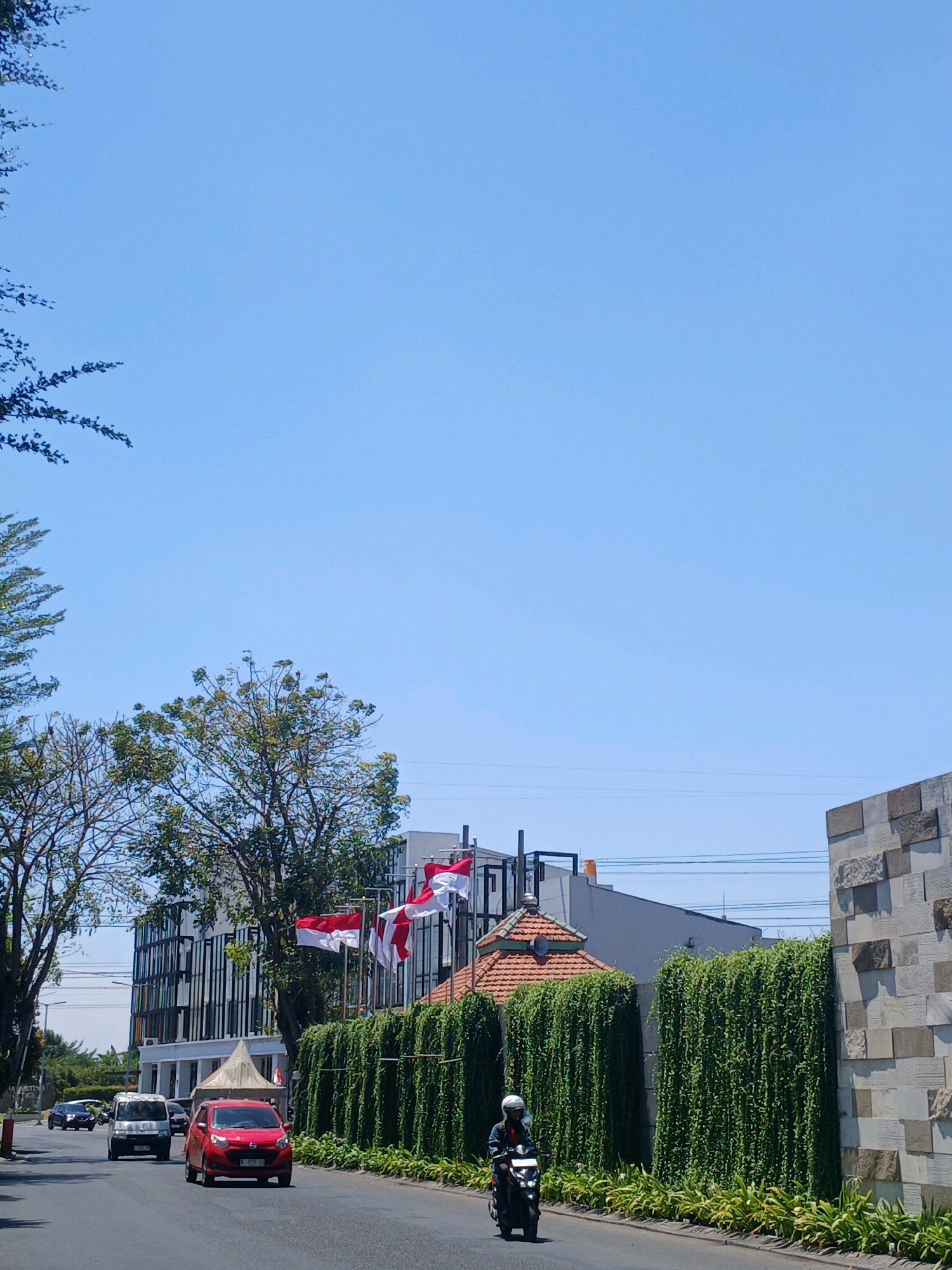 This image captures a street scene in Surabaya, Indonesia, under a clear blue sky. The road is asphalt, with a slight curve to the right. A red car is driving on the road, and a motorcycle with a rider is also visible. On the right side of the road, there is a wall made of textured stone blocks, and a long, vertical garden of green plants stretches along the property line. In the background, a modern building with a dark, patterned facade stands behind a section of red-tiled roof. Indonesian flags are prominently displayed on poles in front of the building. Trees with green foliage are present on the left side of the road, with some branches extending into the upper left corner of the frame. The overall impression is a sunny day in an urban environment with elements of nature and modern architecture.