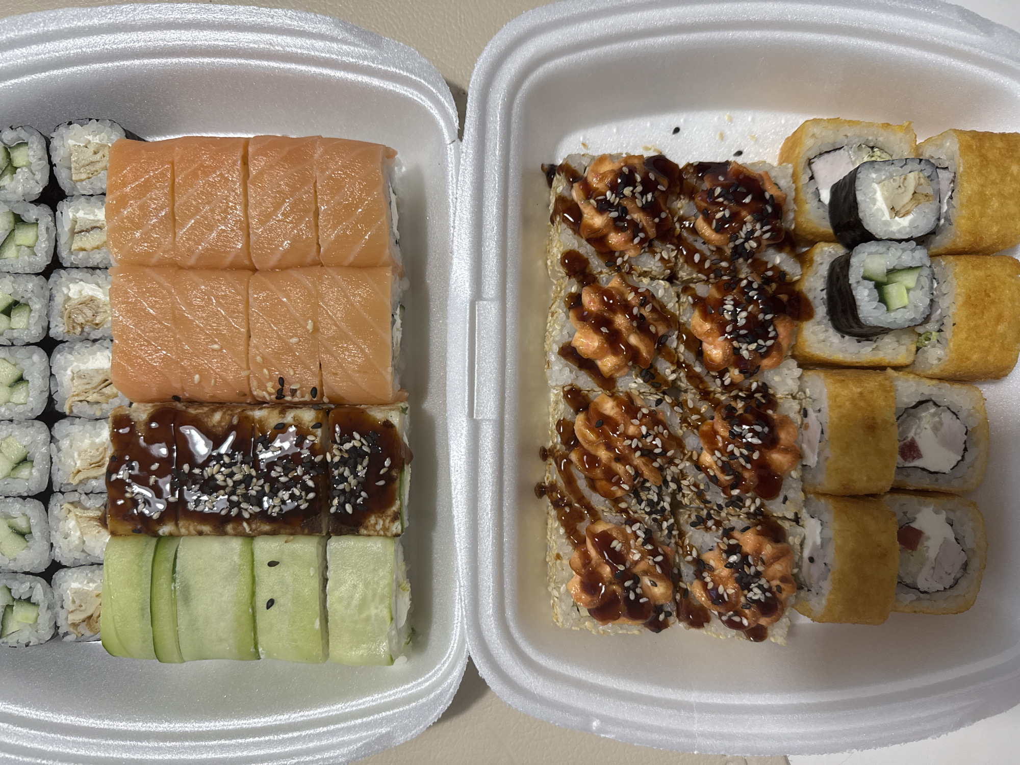 Two white styrofoam containers, likely takeout sushi, are arranged side-by-side on a light beige surface. The container on the left contains rows of sushi rolls, including cucumber rolls, rolls with what appears to be omelet and cream cheese, salmon rolls, and rolls with eel sauce and sesame seeds. The container on the right holds sushi rolls with spicy mayo and sesame seeds on top, some with a crispy outer layer, and other rolls with cream cheese and what appears to be chicken. There is no indication of location within Chaikovsky, Russia. No people are visible in the image.