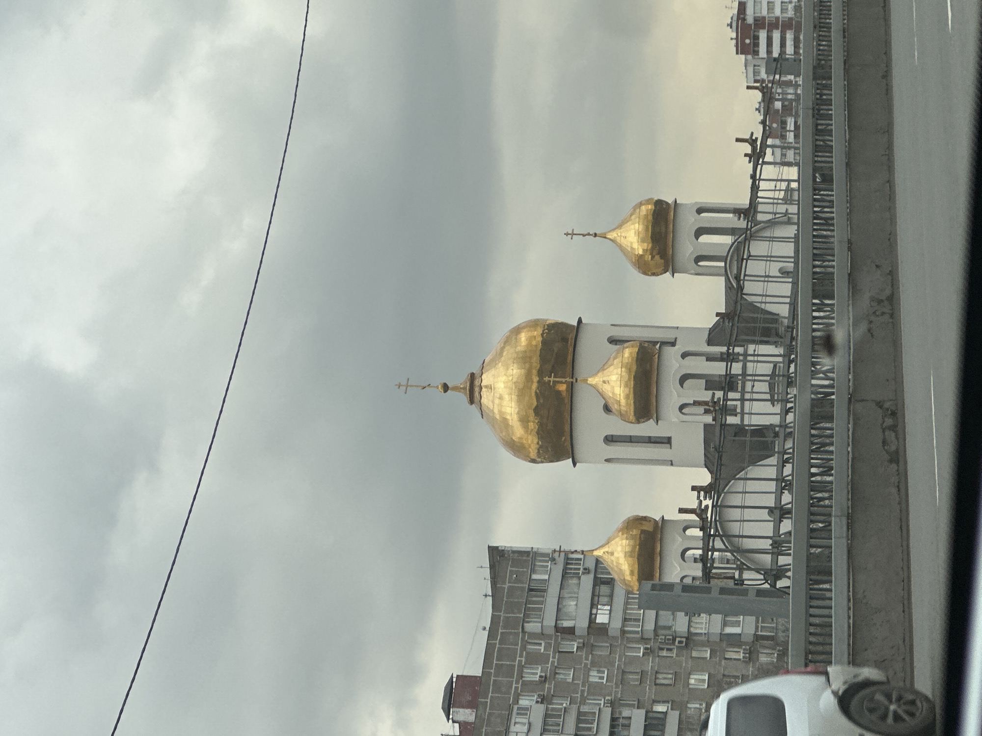 The image depicts a Russian Orthodox church with multiple golden domes against a cloudy sky. The church has white walls with arched windows. It appears to be located in an urban area with residential buildings visible in the background. A bridge or elevated road runs alongside the church, partially obstructing the view. A power line is visible in the upper-left corner. The image was likely taken from inside a moving vehicle. It is difficult to pinpoint the exact location within Moscow based on the provided details.