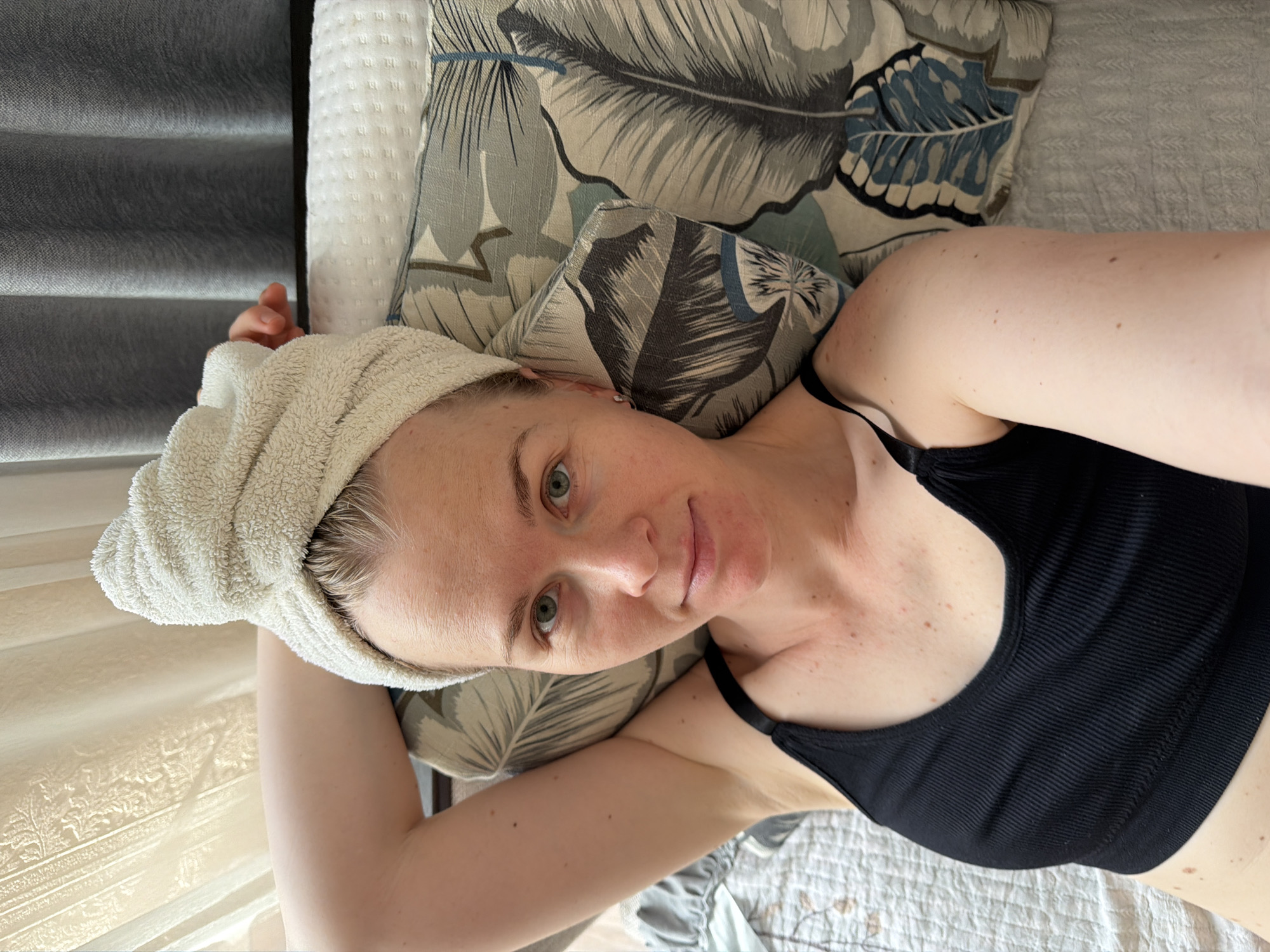 A woman with blonde hair wrapped in a white towel is lying down with her head on a pillow. She is wearing a black tank top and looking at the camera. The scene appears to be indoors, possibly a bedroom, with soft curtains in the background. The lighting suggests it is daytime. The woman has light-colored skin and green eyes. There are some small red marks on her upper chest and shoulder. The pillow has a pattern of large leaves in shades of blue and gray. The overall impression is one of a relaxed, casual moment. There is no indication of the location being in the city of Khabarovsk, Russia.