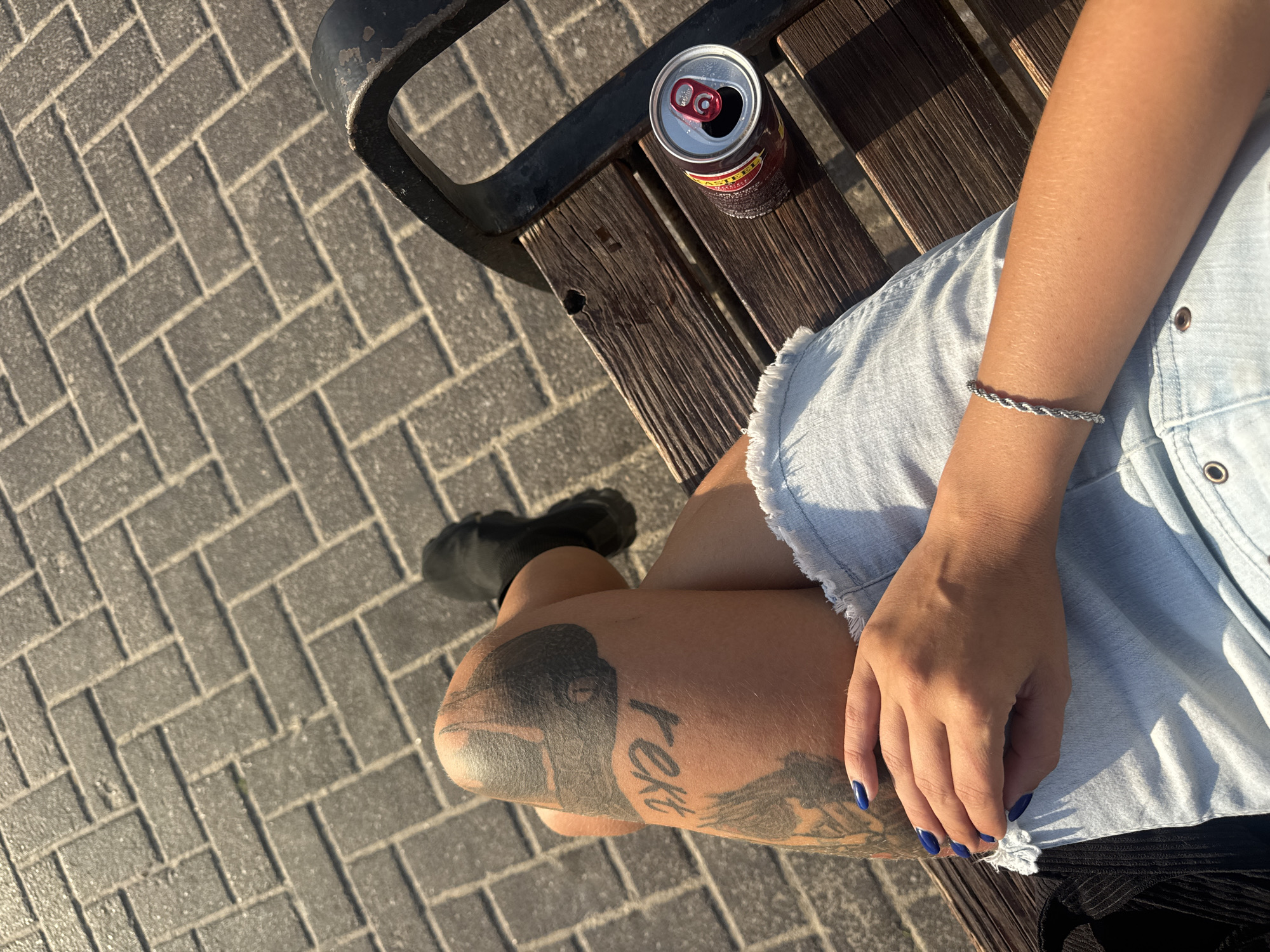 A woman is sitting on a wooden bench with a can of soda next to her. She is wearing a denim skirt and black shoes. The bench is on a paved area with a herringbone pattern. The woman has tattoos on her leg and blue nail polish on her fingernails.