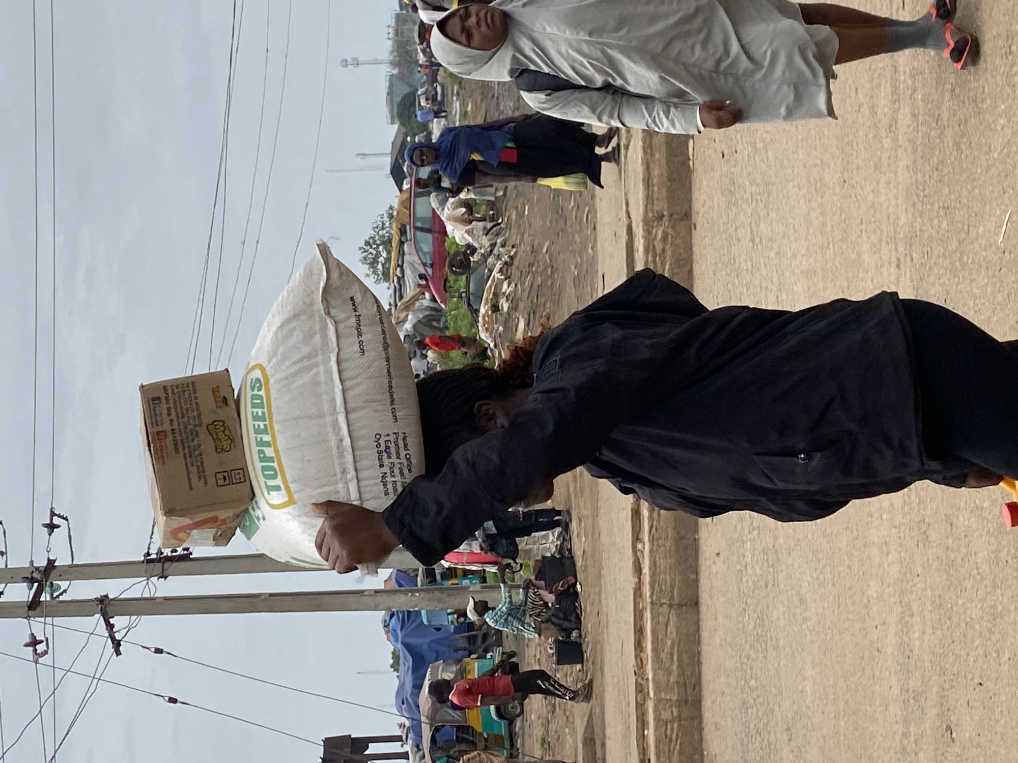 This media file depicts a scene in Bukuru, Nigeria, likely an outdoor market or roadside area. The primary subject is a person carrying a large sack of "TOPFEEDS" and a cardboard box on their head, showcasing a common method of transporting goods. The sack has text indicating "Premier Feed" and an address: "1 Eagle Flour road, Oyo State, Nigeria," along with a website, "www.Imnpic.com". Another person, wearing a grey hooded garment, is visible in the background, looking towards the camera.
The setting appears to be a somewhat cluttered environment with various objects and debris on the ground, suggesting a busy and perhaps unkempt area. There are power lines and poles in the foreground, indicating urban infrastructure. Other people and what look like vehicles or stalls are present in the background, contributing to the impression of a lively, active marketplace. The lighting suggests it could be daytime, possibly overcast, given the muted tones. No specific emotions are overtly conveyed by the individuals in the frame. The overall impression is one of daily life and commerce in a Nigerian location.