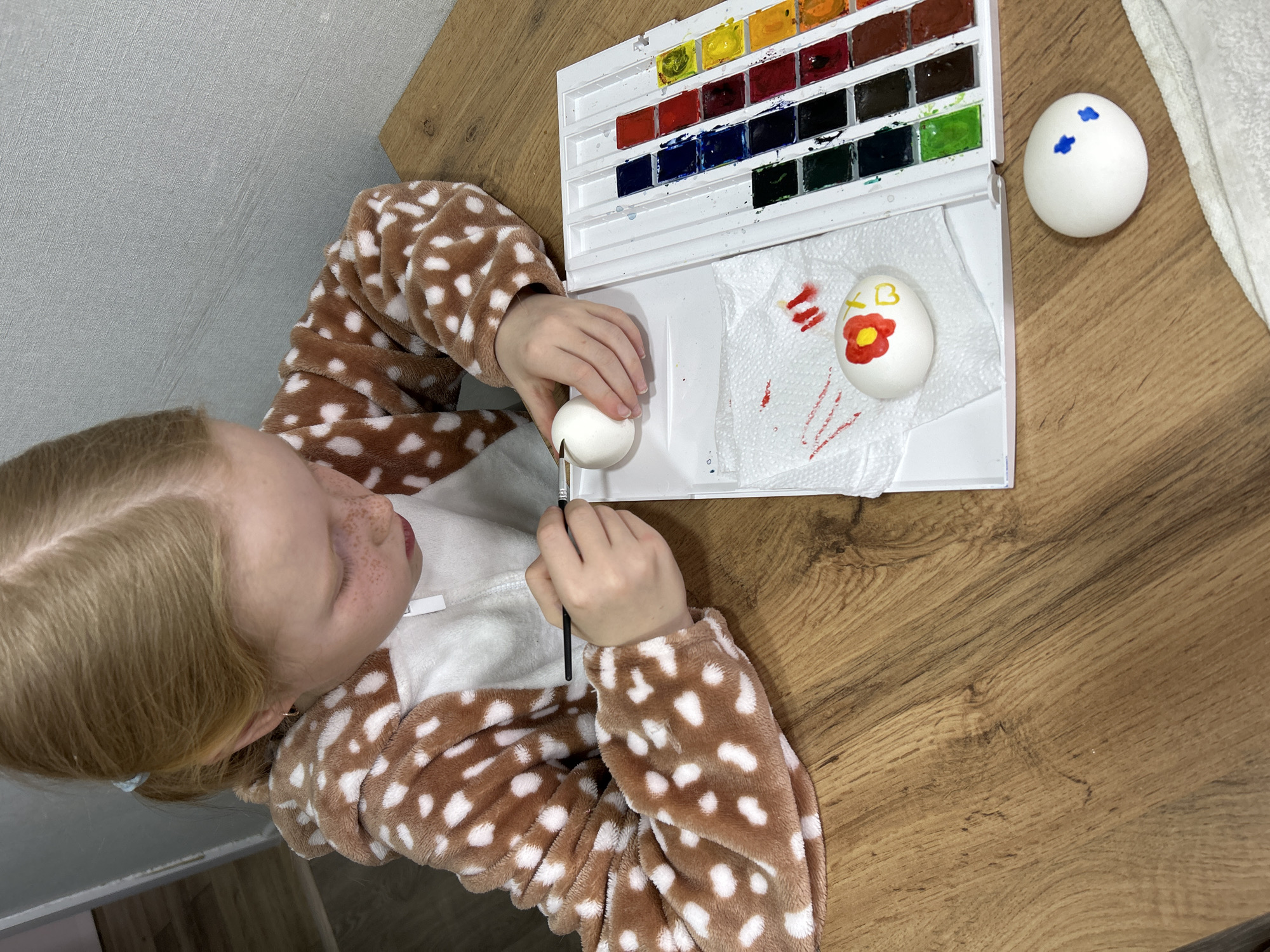 A high-angle indoor shot shows a young girl in a brown and white spotted onesie painting Easter eggs at a table. The girl, with blonde hair and freckles, is focused on her task. She holds a white egg in her left hand and a paintbrush in her right. Next to her, on a white tray, is a watercolor paint palette with a variety of colors. Two eggs sit on the tray: one is plain white with a few blue dots, and the other has a red flower and the letters "XB" painted on it. The tray rests on a white paper towel. The table is made of wood with a light brown, patterned surface. The scene is lit from above, casting soft shadows. The location is implied to be Chaikovsky, Russia, based on the Russian letters painted on the egg.