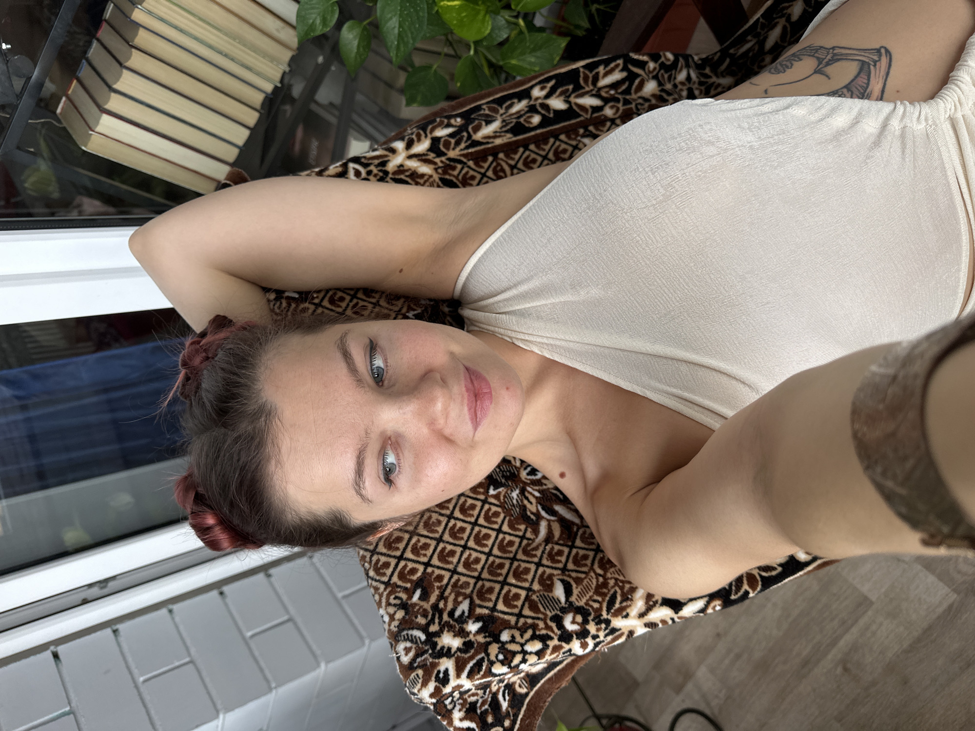 A young woman is captured in a high-angle, close-up selfie, lying down and looking directly at the camera with a soft, pleasant smile. Her head is tilted, and her arms are raised and relaxed behind her head, highlighting a light-colored, possibly cream or off-white, sleeveless top or dress with gathered fabric detailing. She has fair skin, bright blue eyes, and dark reddish-brown hair styled into two small buns or braids on the sides of her head. A distinct, dark-colored illustrative tattoo, featuring what appears to be a skeletal or mechanical design, is visible on her upper left arm.
She is resting on a patterned blanket or rug adorned with intricate brown, black, and white geometric and floral designs. In the background, to her left, a neat stack of books with light-colored pages is visible near a window or glass partition. A vibrant green-leafed houseplant is also present, partially seen behind the books. To her right, a segment of a light-colored wooden floor is in view. The scene suggests a relaxed, domestic setting, possibly on a balcony or near a large window, illuminated by natural daylight. The image portrays a moment of calm and ease, taken in Vladimir, Russia.