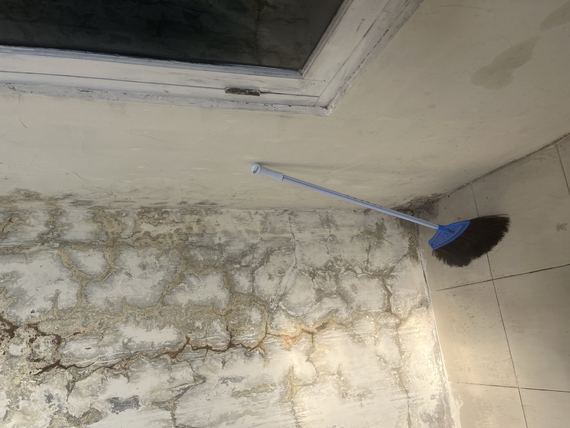 The image depicts an interior corner of a room. On the left, the wall shows significant water damage and staining, with a textured, peeling surface and brown and grey discoloration forming an intricate, web-like pattern. The floor is tiled with square, light-colored tiles. A blue broom with dark bristles rests against the right wall, leaning in the corner between the damaged wall and a plain, cream-colored wall. This cream-colored wall has visible cracks and some water spots. To the right, a window frame, painted white, is visible. The window itself is dark, suggesting it may be closed or its glass is dark. The overall scene suggests a poorly maintained or recently damaged interior space. The location is identified as Cirahayu, Indonesia, though there are no specific visual cues within the image to confirm this location. There are no people present in the image, and no actions are being depicted beyond the static placement of the broom.