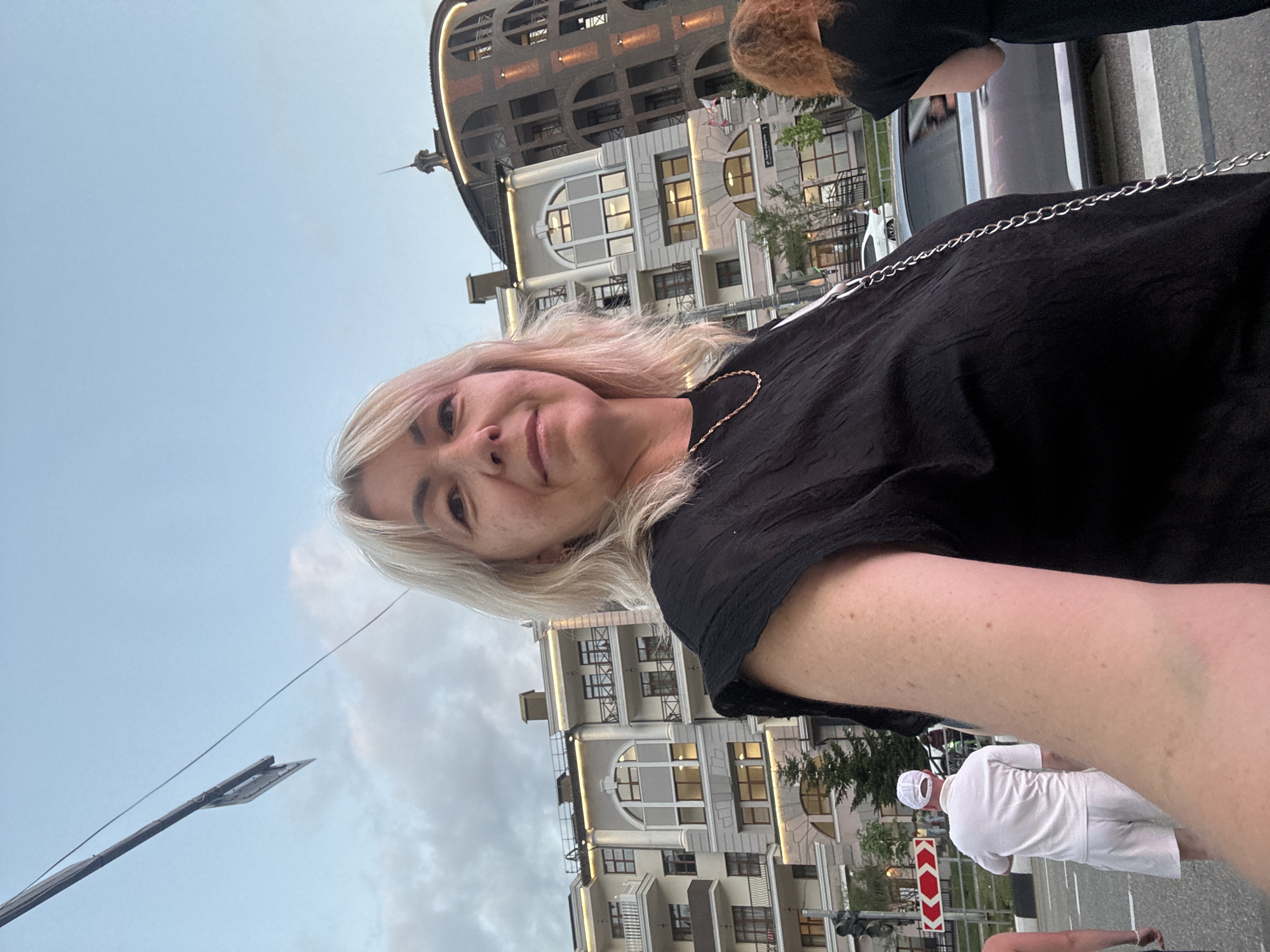 A woman with blonde hair and a black top stands in front of buildings in Krasnaya Polyana, Russia. She is facing the camera and smiling. In the background, there are several buildings, including a multi-story apartment complex and a round, brick building with lights. A street with cars and pedestrians is visible in front of the buildings. A traffic sign with red arrows is also present. The sky is cloudy and overcast.