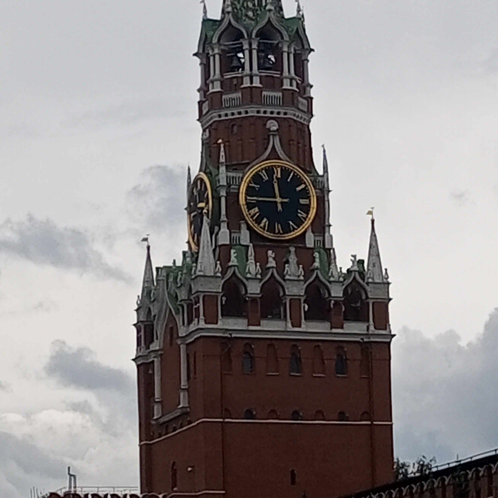 The image is a close-up, low-angle shot of the Spasskaya Tower in Moscow, Russia. The tower is made of red brick and has several clocks. The main clock face, prominently displayed, shows the time as approximately 4:53. Another clock face is partially visible to the left. The tower is topped with green spires and decorative elements. The sky is overcast with gray clouds.