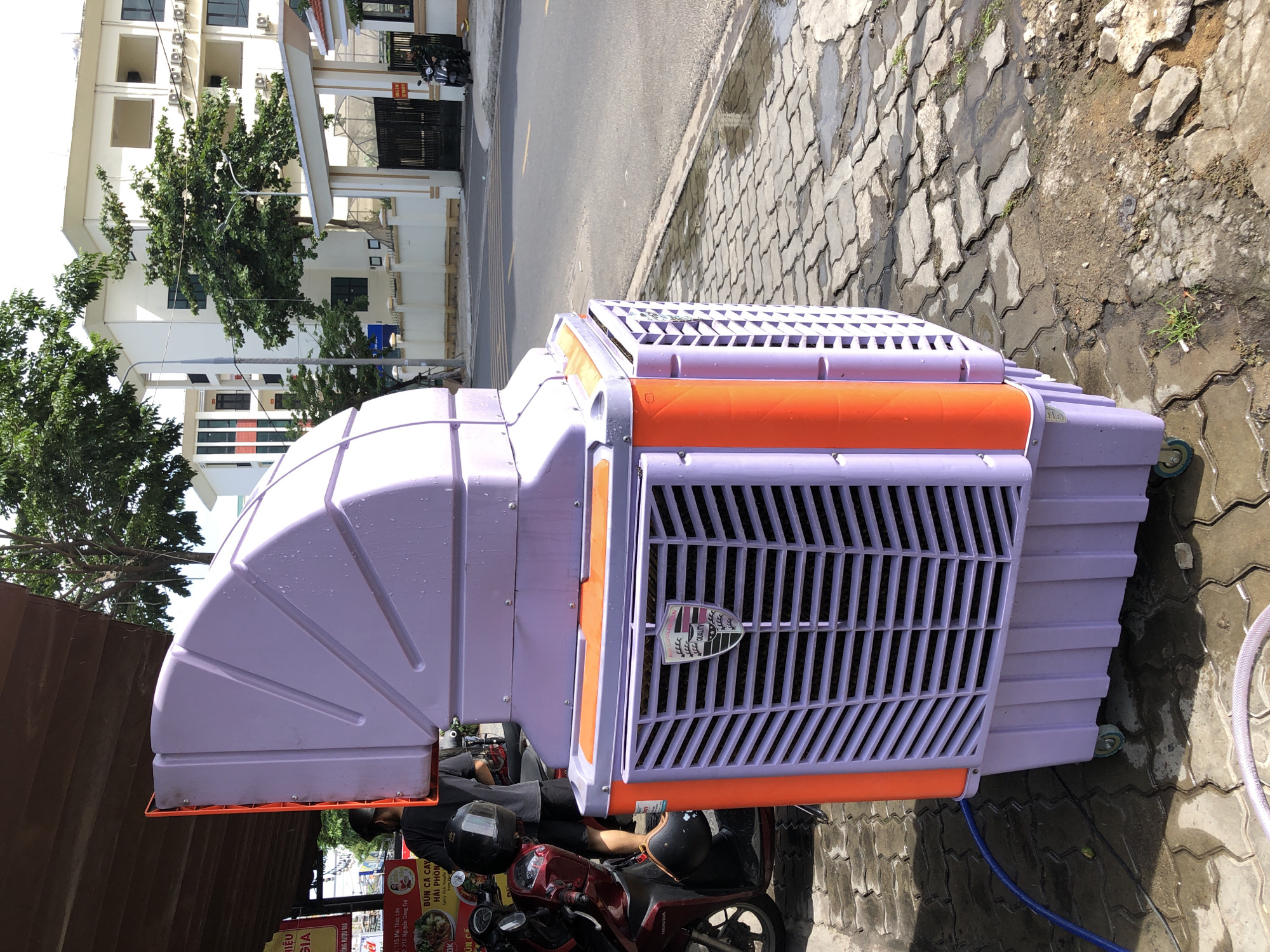 In the city of Đà Nẵng, Vietnam, on a paved outdoor area adjacent to a street, a large, lavender-colored evaporative cooler is positioned. The cooler has orange accents on its sides and a prominent, angled top section with a fan grille. A blue hose is connected to the cooler and lies on the wet pavement. To the left of the cooler, a red motorcycle is parked, with a person wearing a dark shirt and shorts standing next to it. In the background, a white building with multiple stories and a gated entrance is visible, along with trees and streetlights. The ground is a mixture of paving stones and soil, with puddles of water indicating recent moisture.